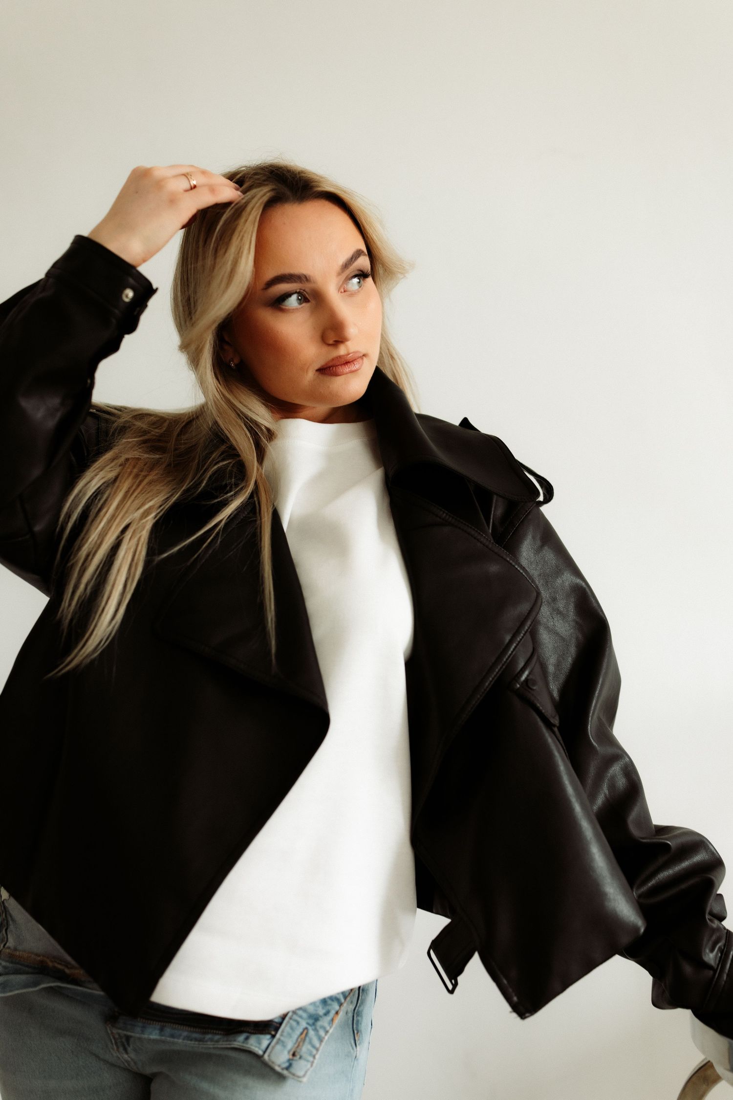 Woman in black leather jacket and white turtleneck with jeans poses against white background.