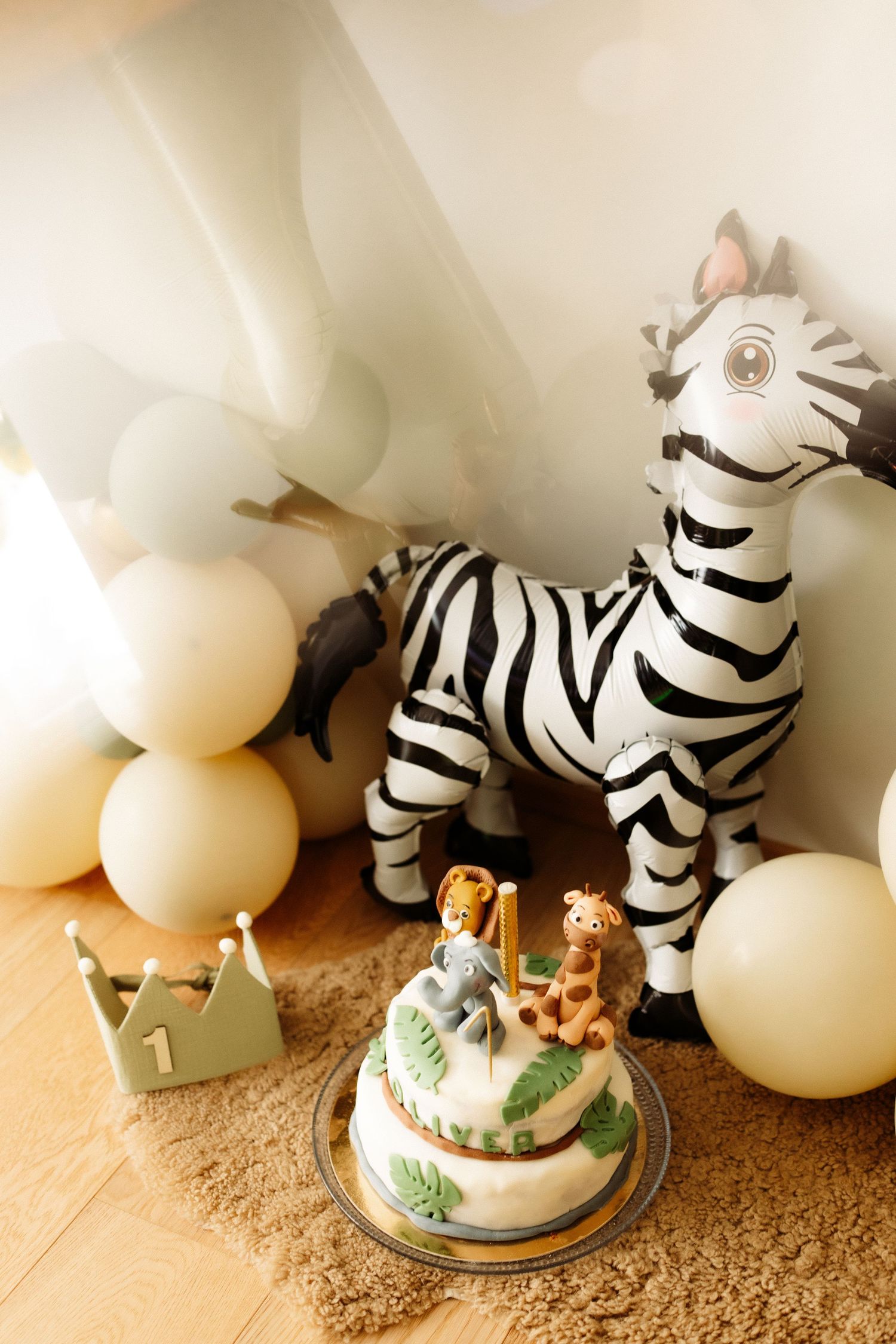 A safari-themed first birthday cake with zebra print decorations and white balloons arranged on a beige carpet.