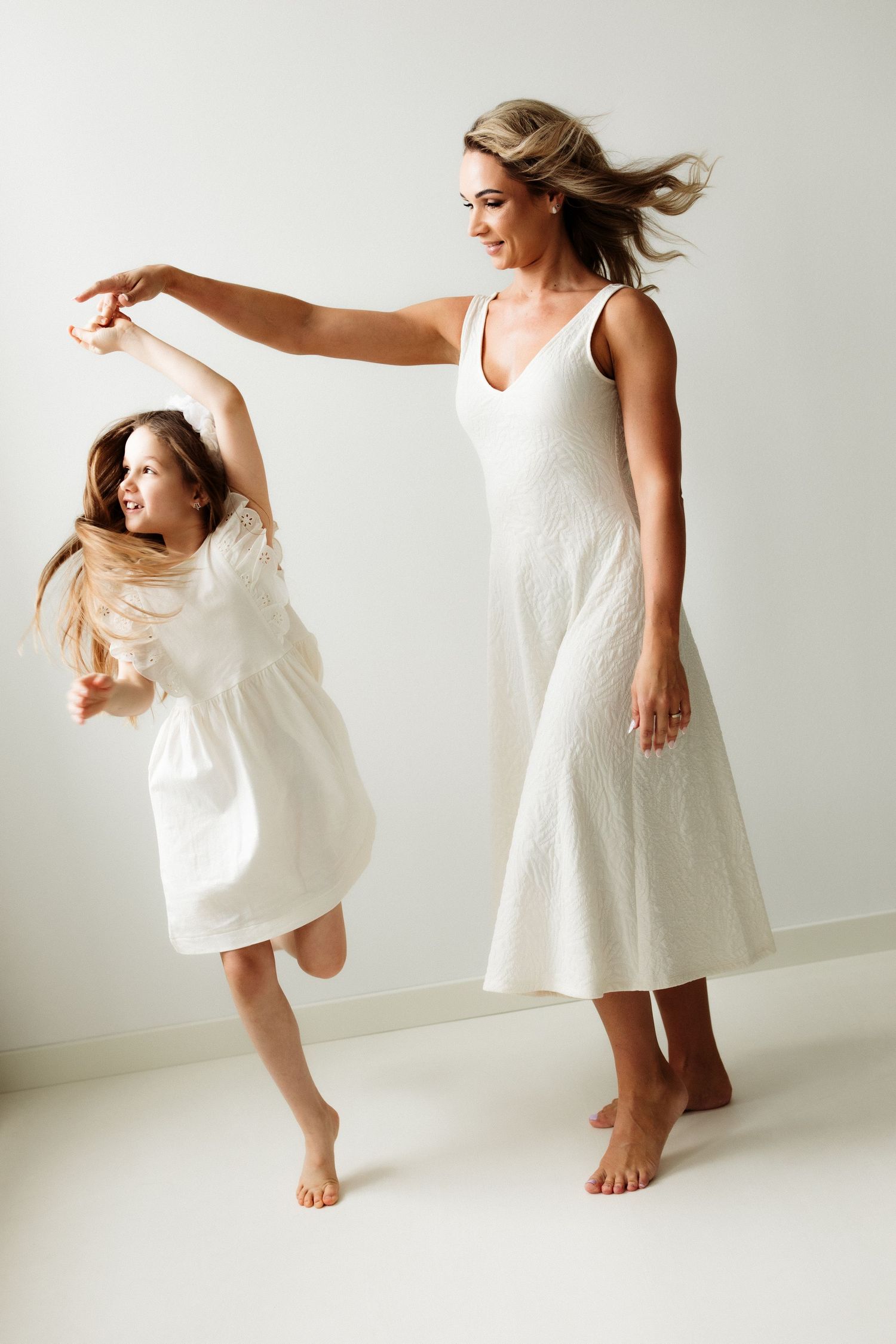 A woman and young girl in matching white dresses dance together barefoot, their hair flowing gracefully.