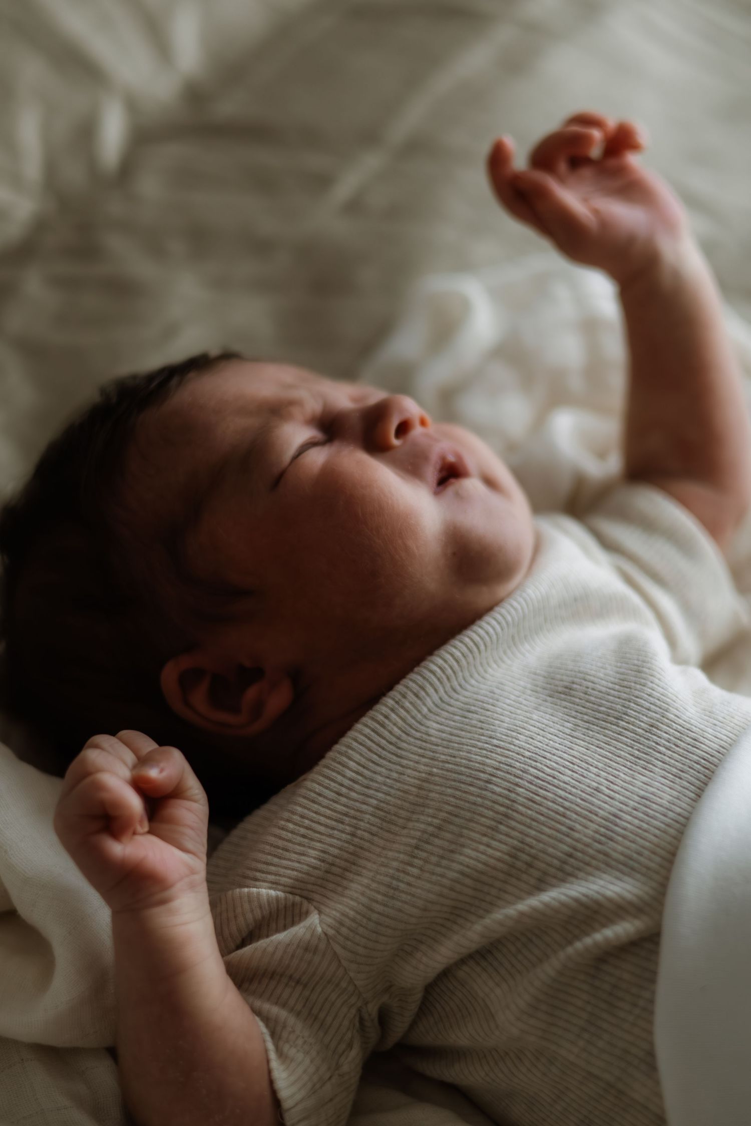 A peaceful newborn baby sleeping on a white bed, wearing a cream knit outfit with tiny fists raised.