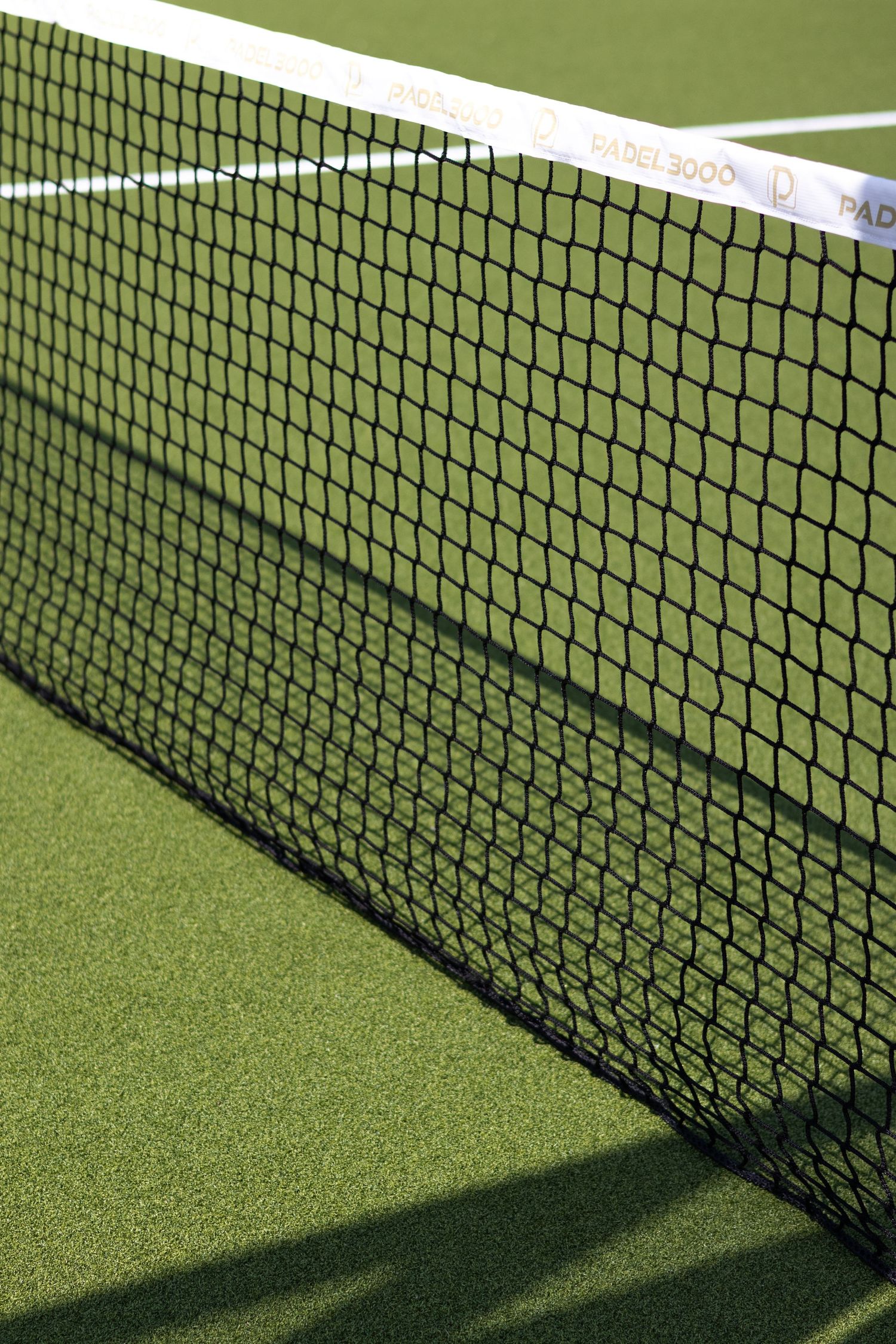 Tennis court net casting shadows on bright green synthetic turf in afternoon sunlight.