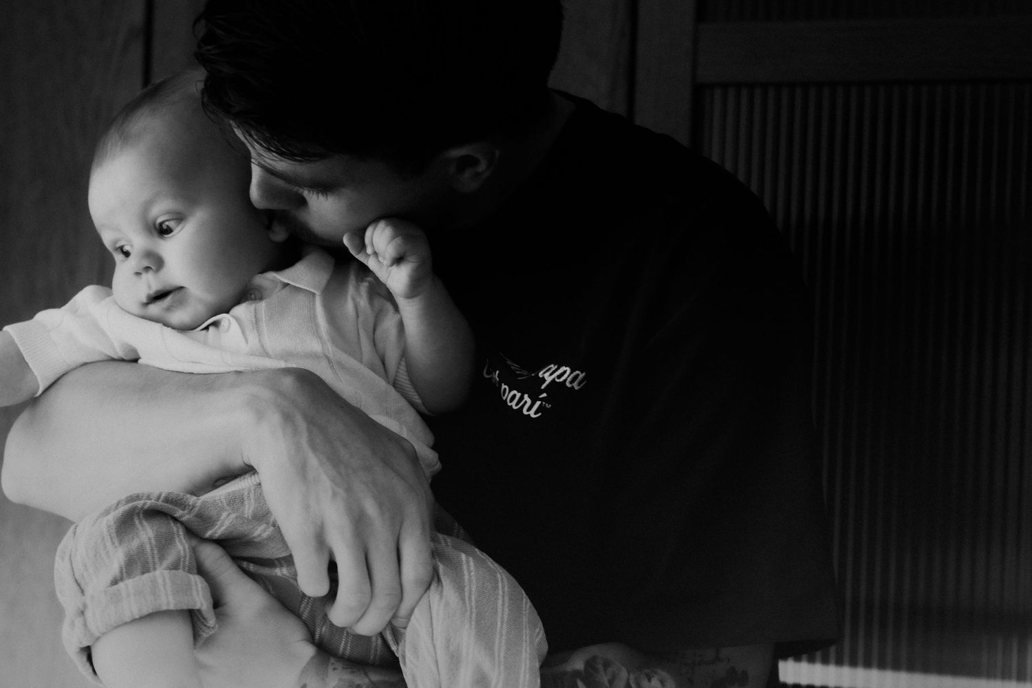 Black and white photo of a parent tenderly kissing a newborn baby on the head while cradling them gently.