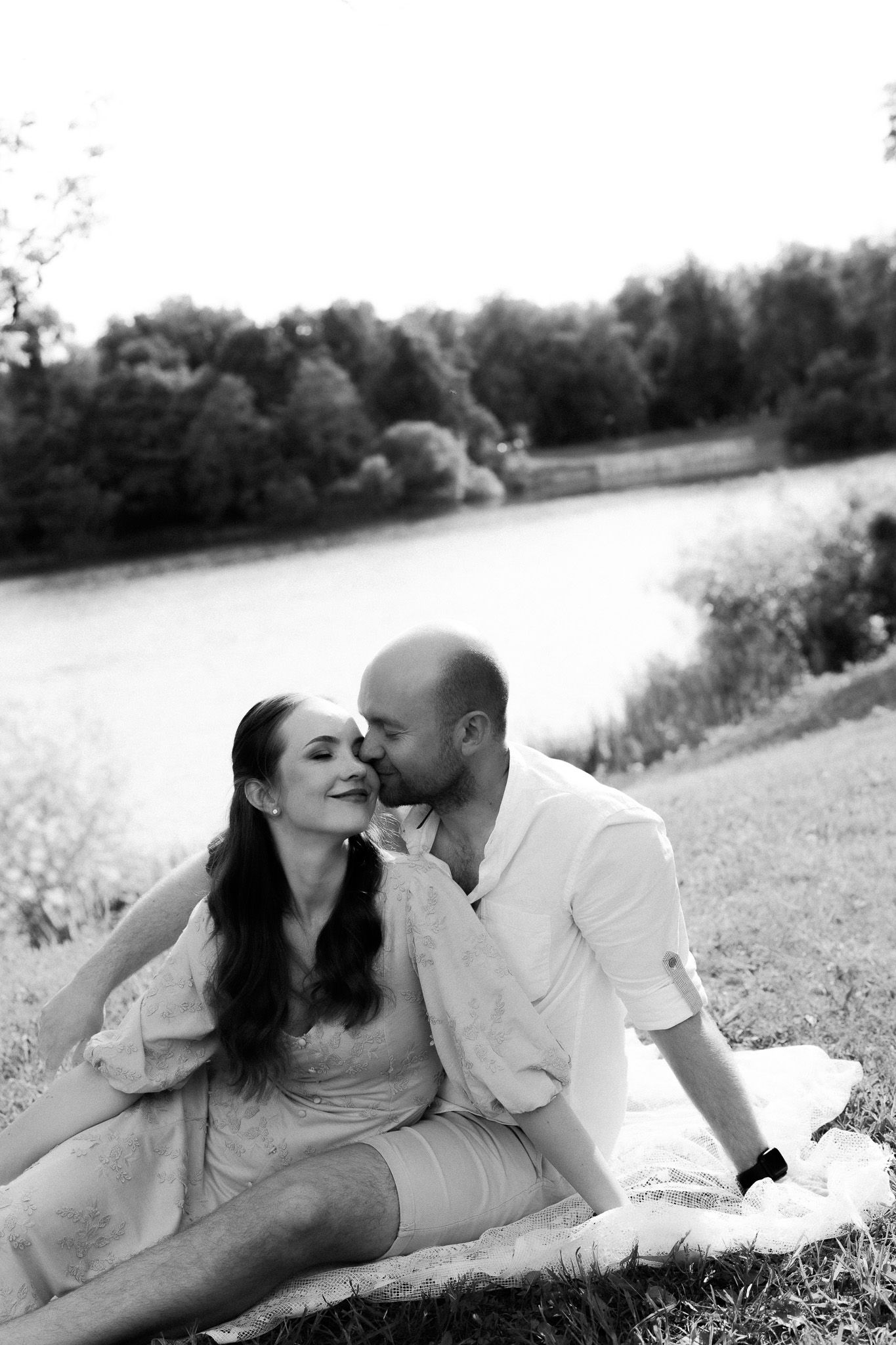 Black and white photo of a couple sitting by a lake, man kissing woman on cheek during a romantic outdoor picnic.