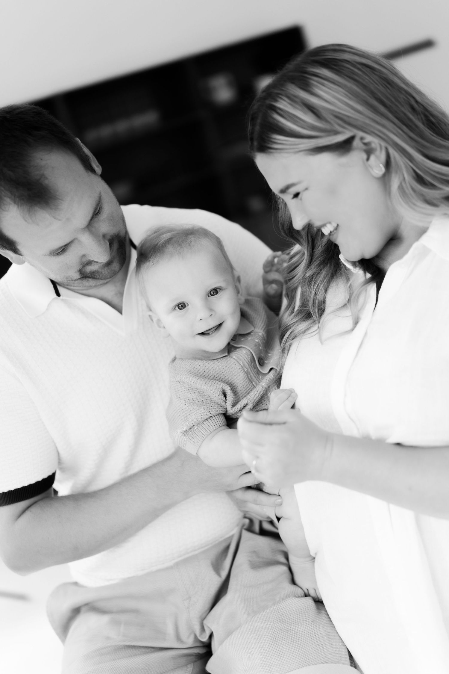 Black and white photo of happy parents holding their smiling baby together indoors.