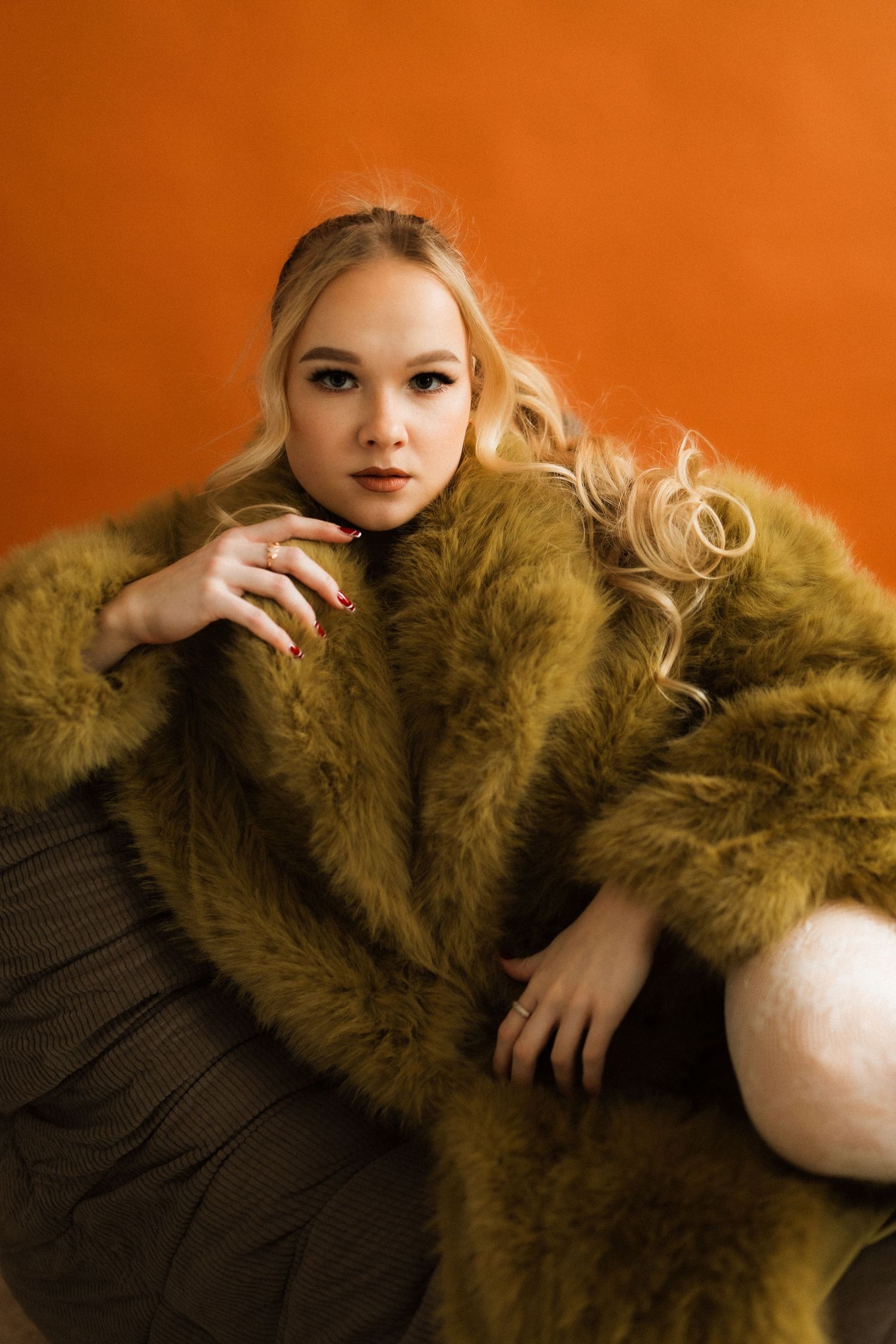 Fashion portrait against orange background wearing olive faux fur coat and dark dress in artistic poses.