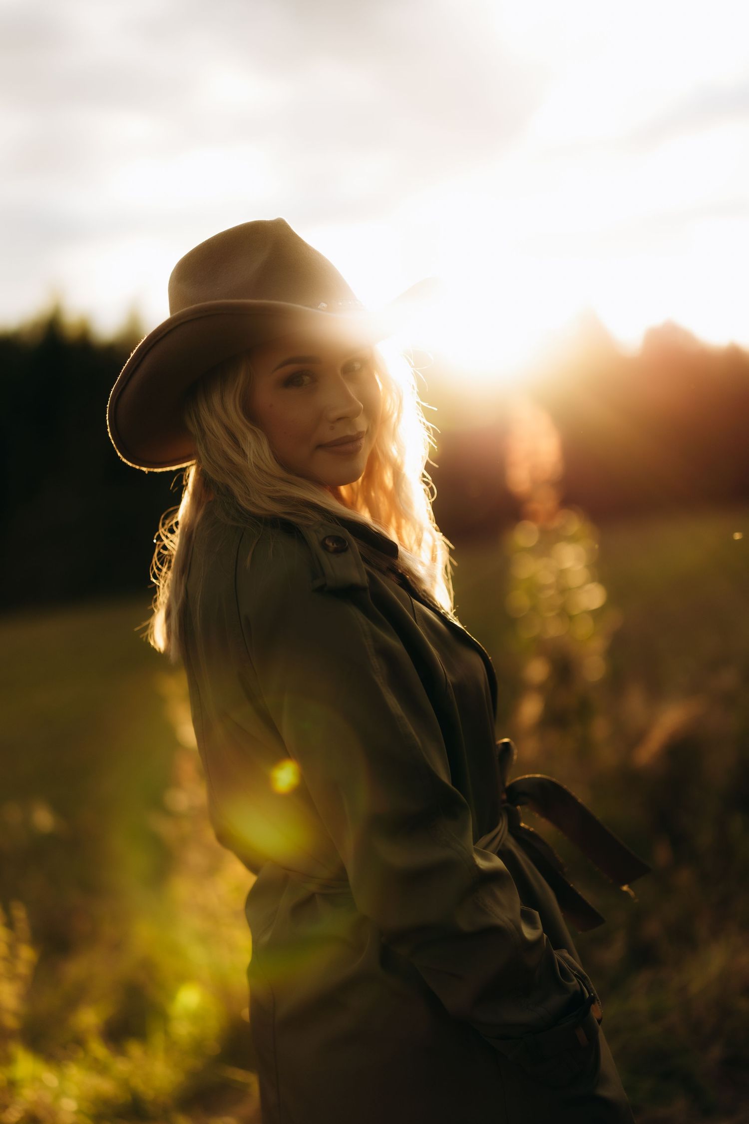 A blonde woman in a wide-brim hat and olive trench coat poses in a sunlit field at golden hour.