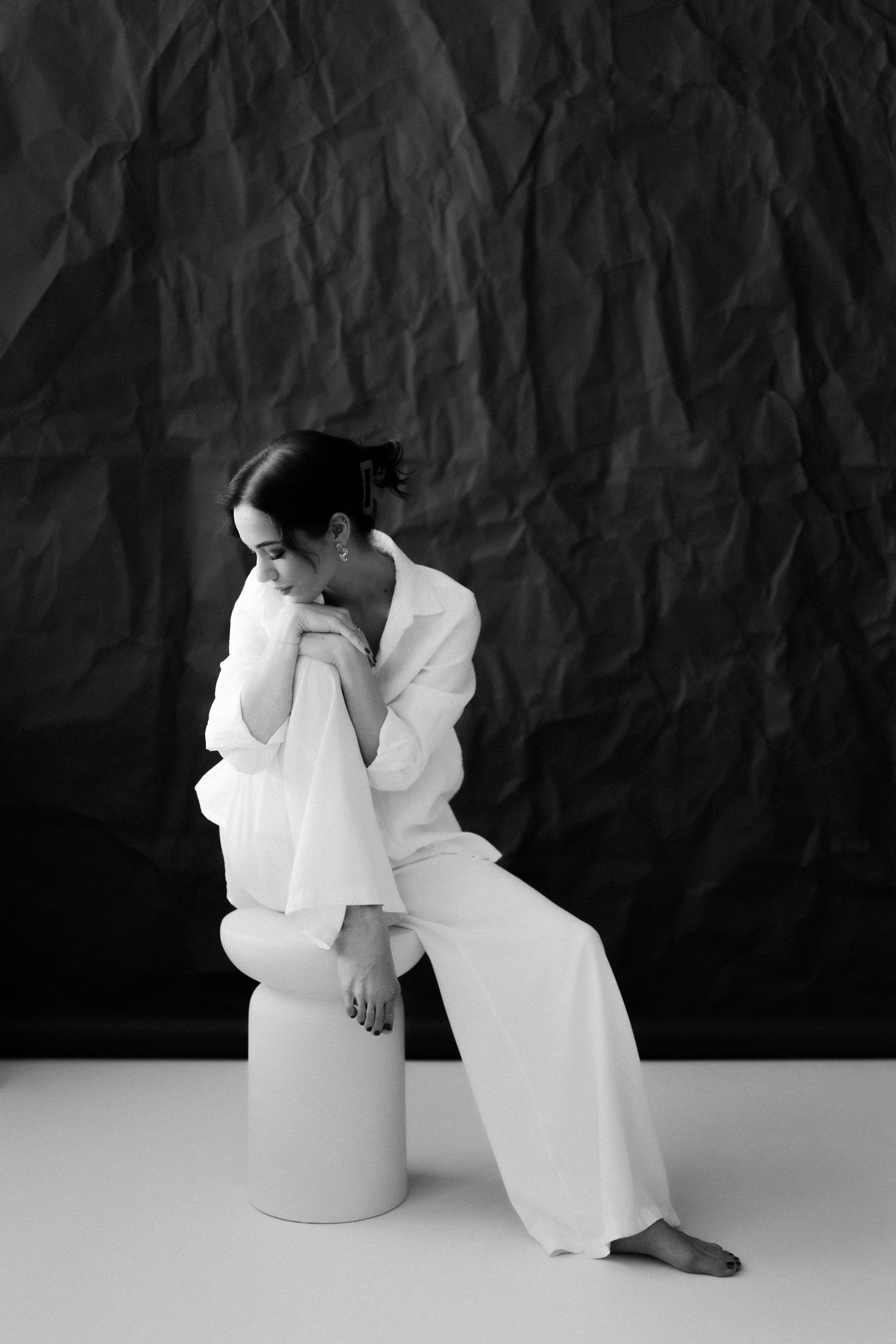 Black and white photo of a woman in white outfit sitting on a cylinder stool, resting her head on her hands.