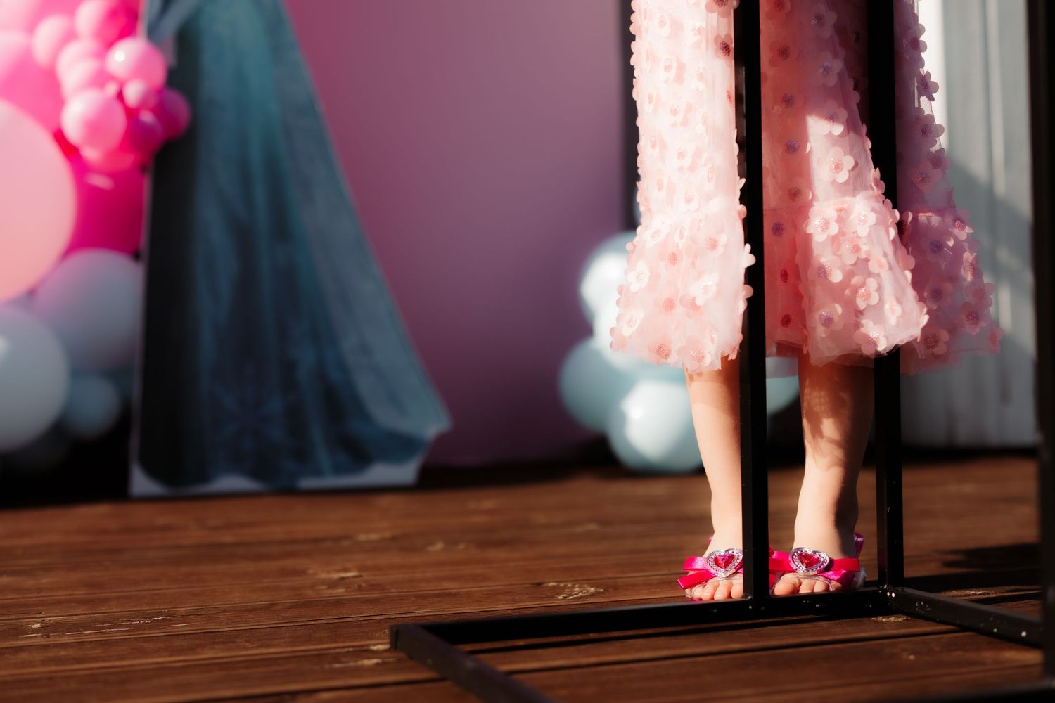 Young girl in pink ruffled dress and floral shoes stands on wooden platform near balloons.