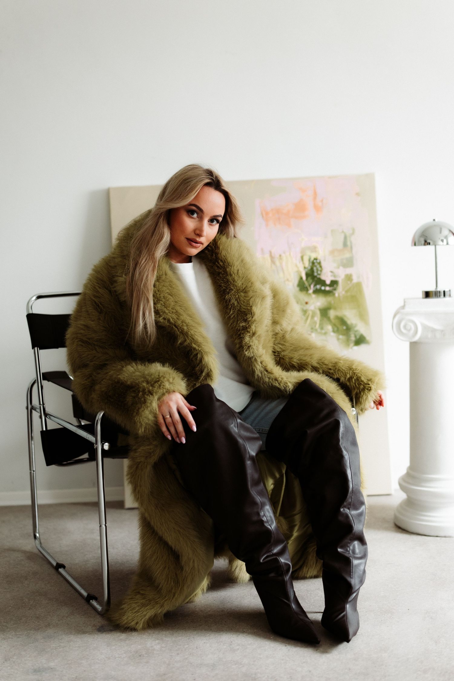Woman in olive fur coat, leather pants, and boots sits on chair before abstract painting.