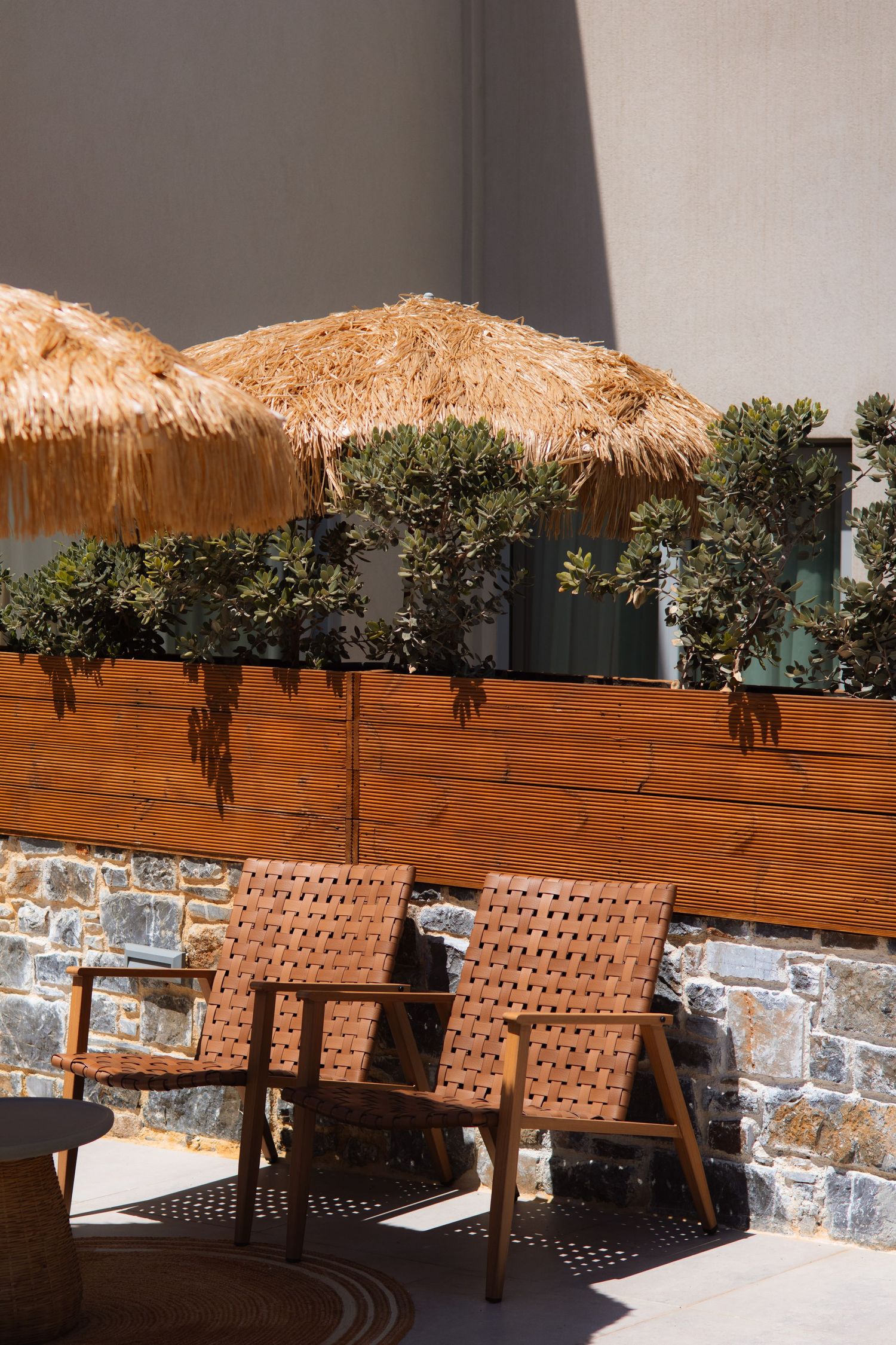 Cozy outdoor seating area with woven leather chairs and thatched umbrellas against a stone and wood wall.