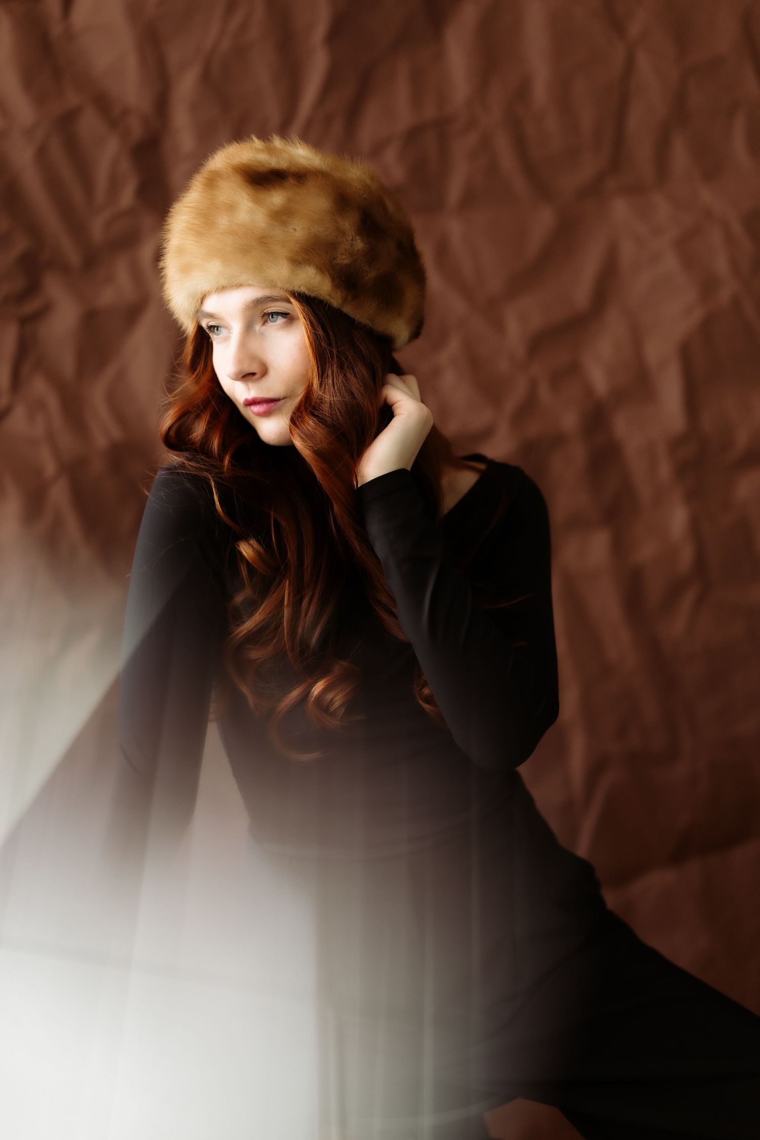 A person in a fur hat poses against a dark brown backdrop in a moody artistic portrait series.