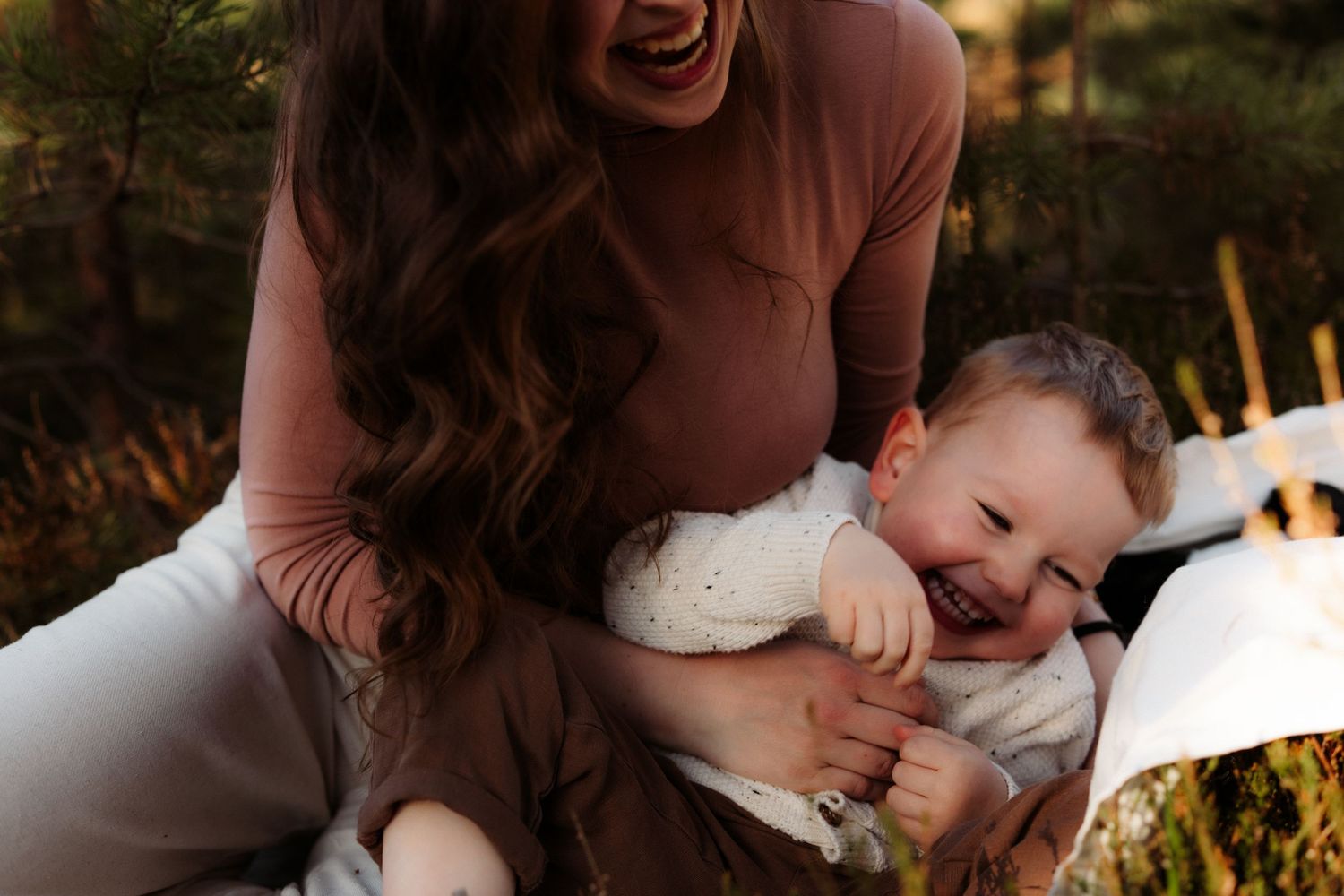 A laughing mother cradles her smiling baby outdoors in warm golden light, sharing a joyful moment together.