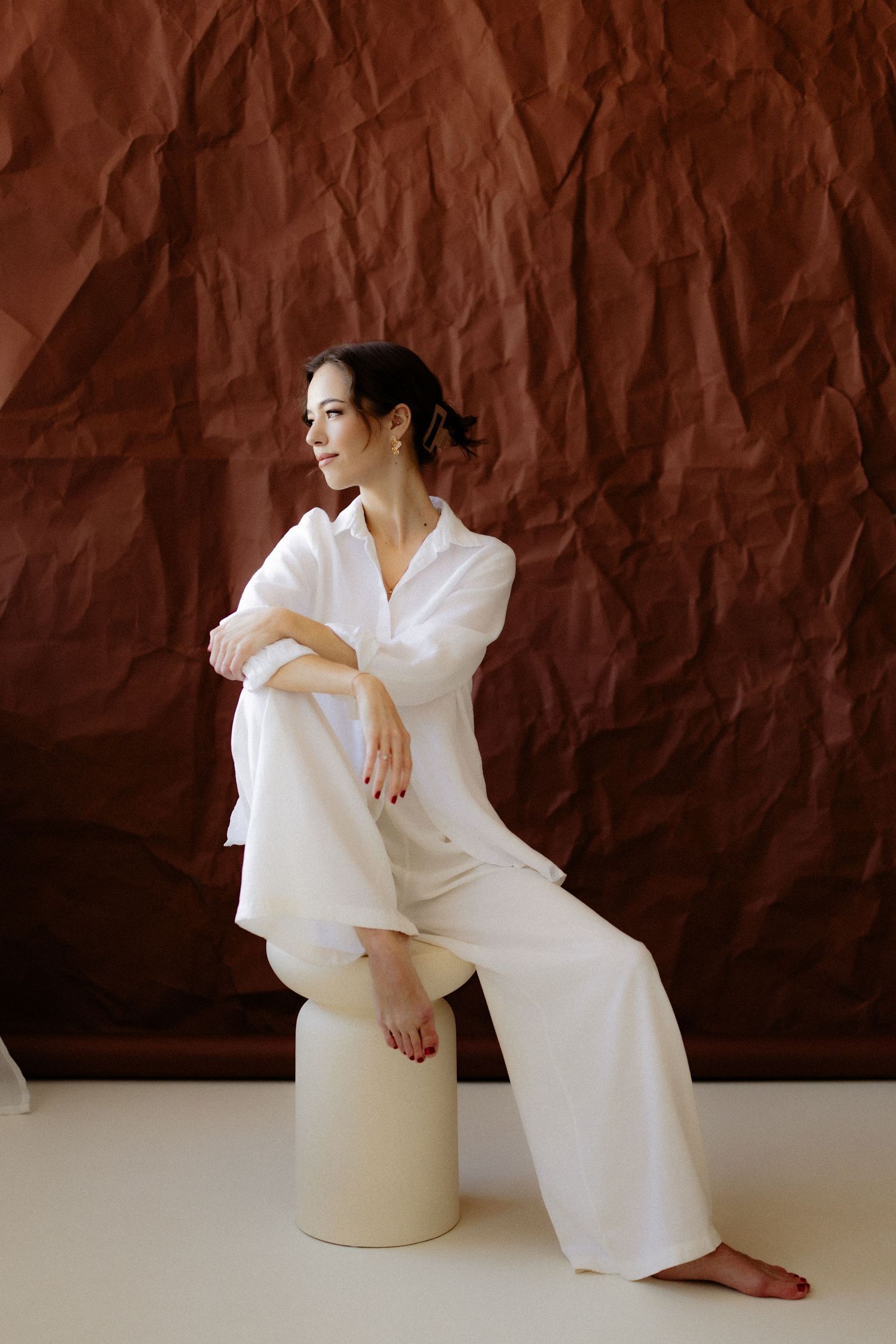 Woman in white linen outfit sitting on a cream stool against a textured brown backdrop in a studio setting.