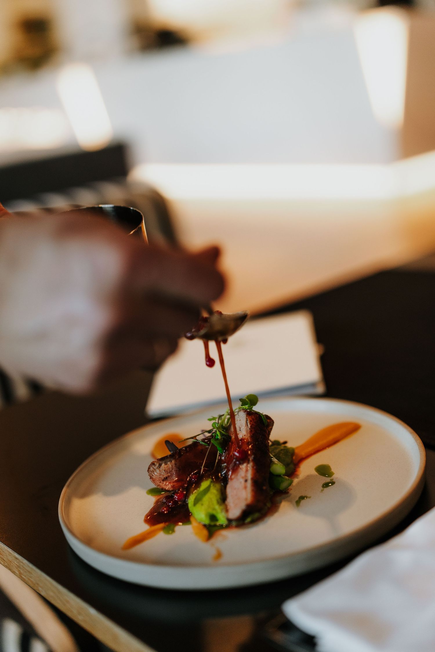Chef pouring dark sauce over elegantly plated dish on white ceramic plate in upscale restaurant setting.