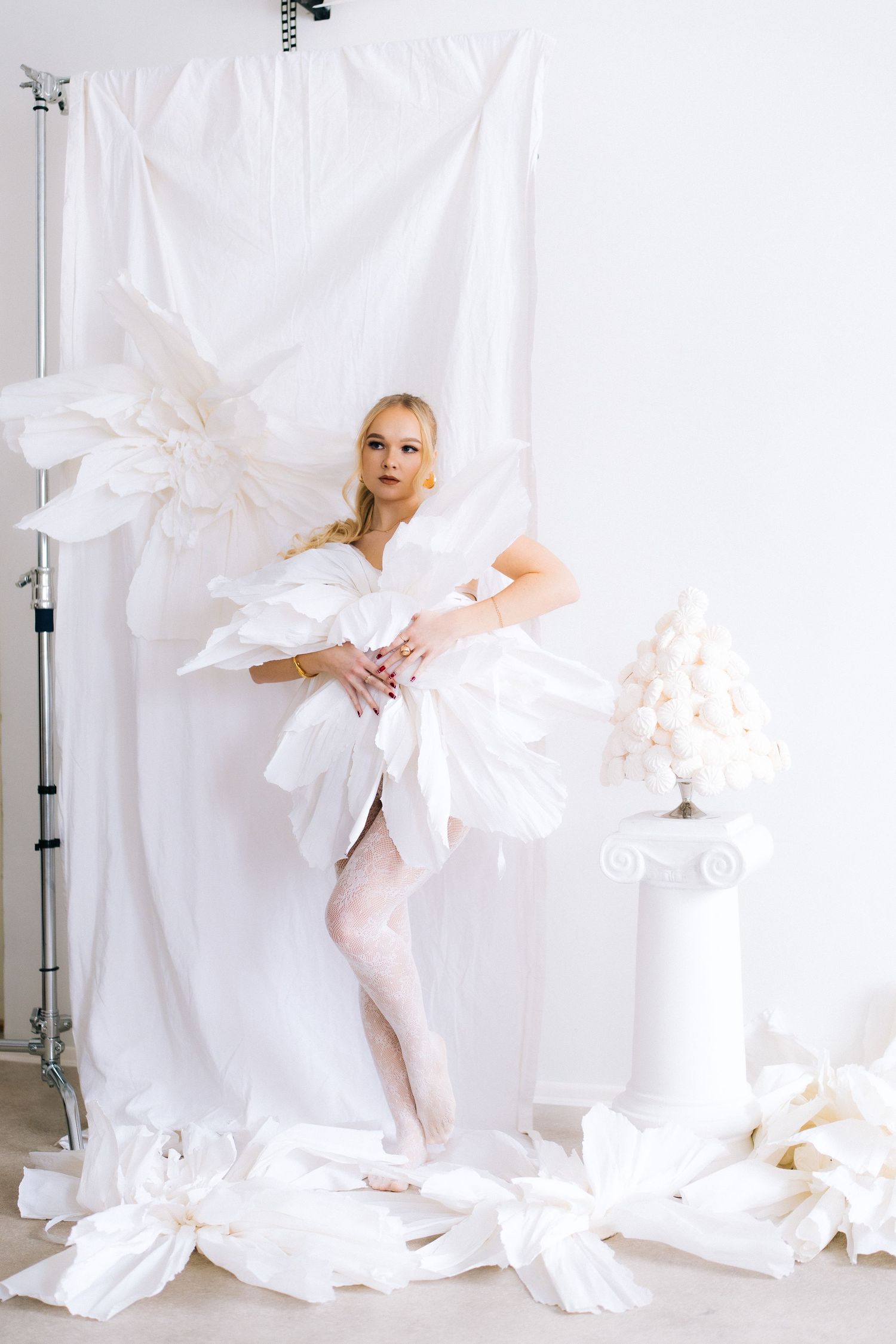 Artistic fashion photo with white paper decorations and flowing fabric against a minimalist white backdrop in a studio setting.