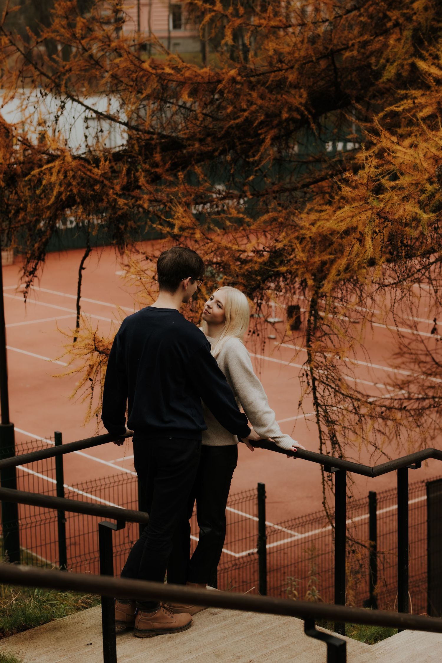 A couple stands together overlooking a tennis court surrounded by autumn trees with orange foliage at sunset.