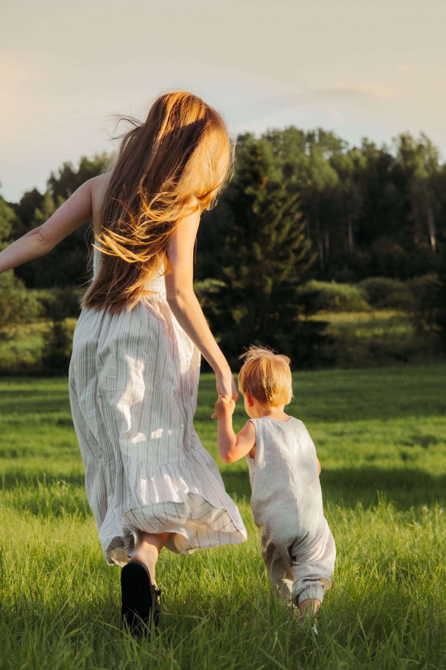 A mother and young child run hand-in-hand through a lush green meadow at golden hour.
