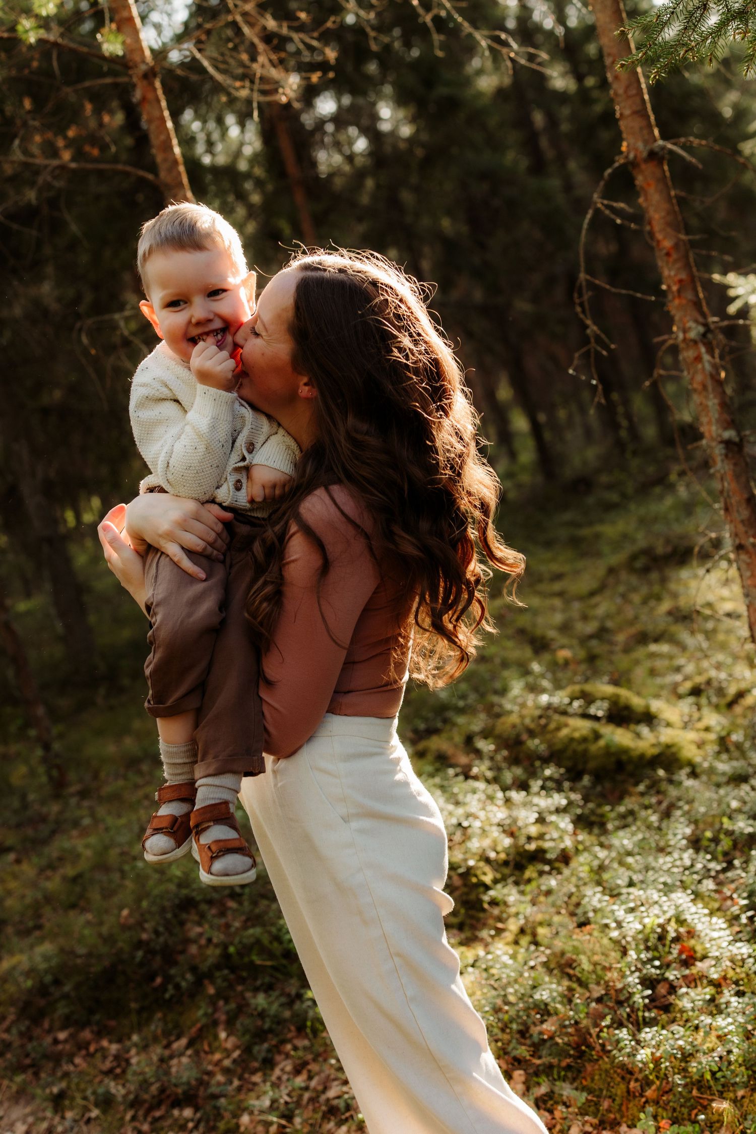 A mother kisses her laughing toddler while holding him in a sunlit forest setting.