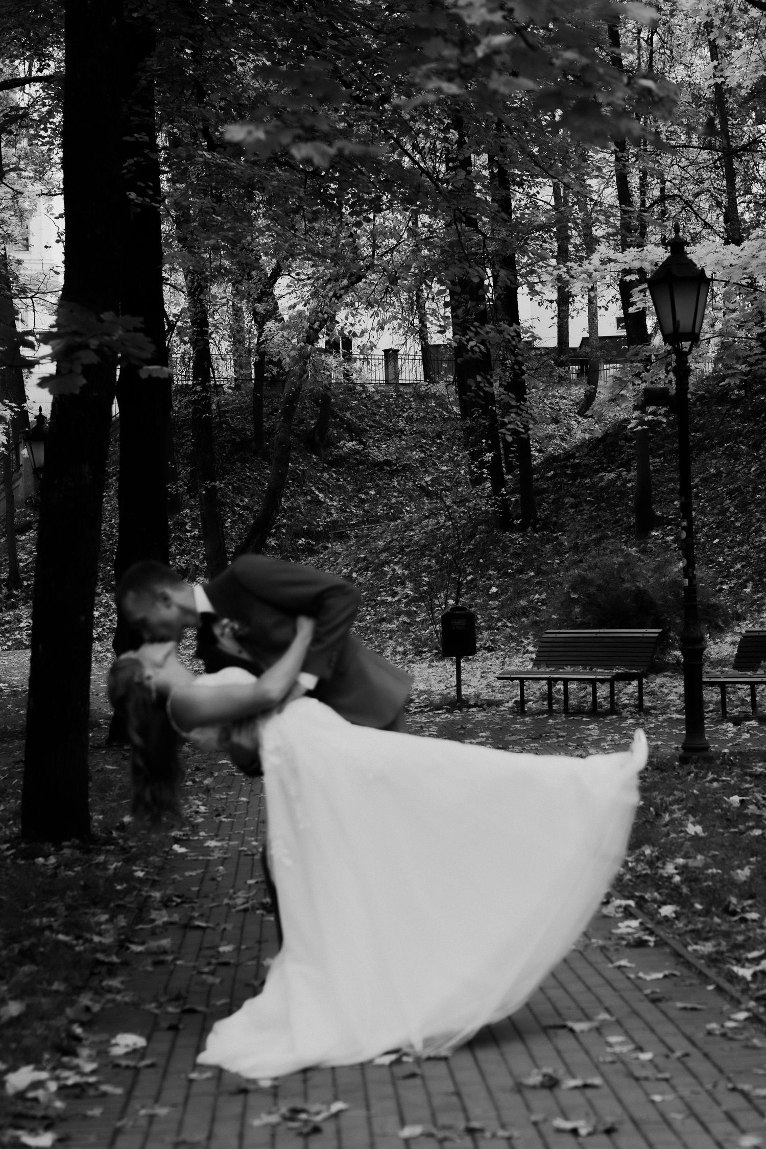 A romantic black and white photo of a couple dancing in a wooded park setting with a vintage lamp post.