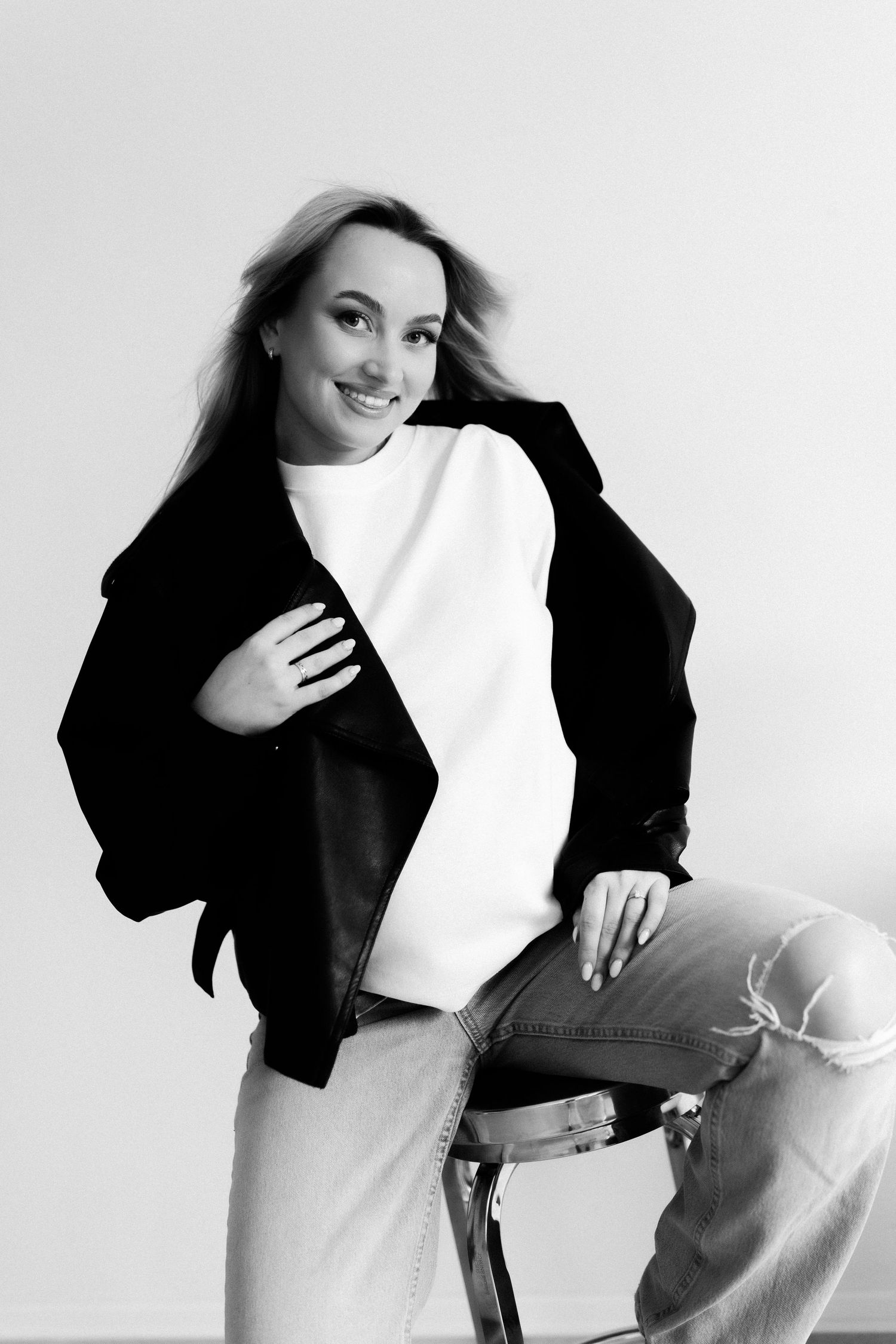 Black and white portrait of a smiling blonde woman in a white top, black jacket, and ripped jeans sitting on a chair.