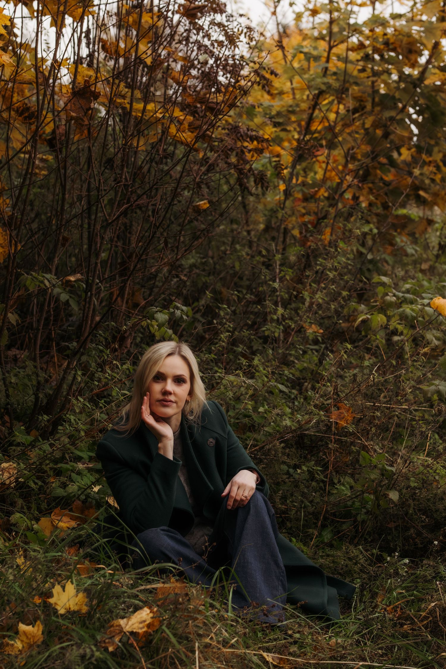 A figure rests among autumn foliage wearing dark clothing in a moody outdoor photoshoot series.