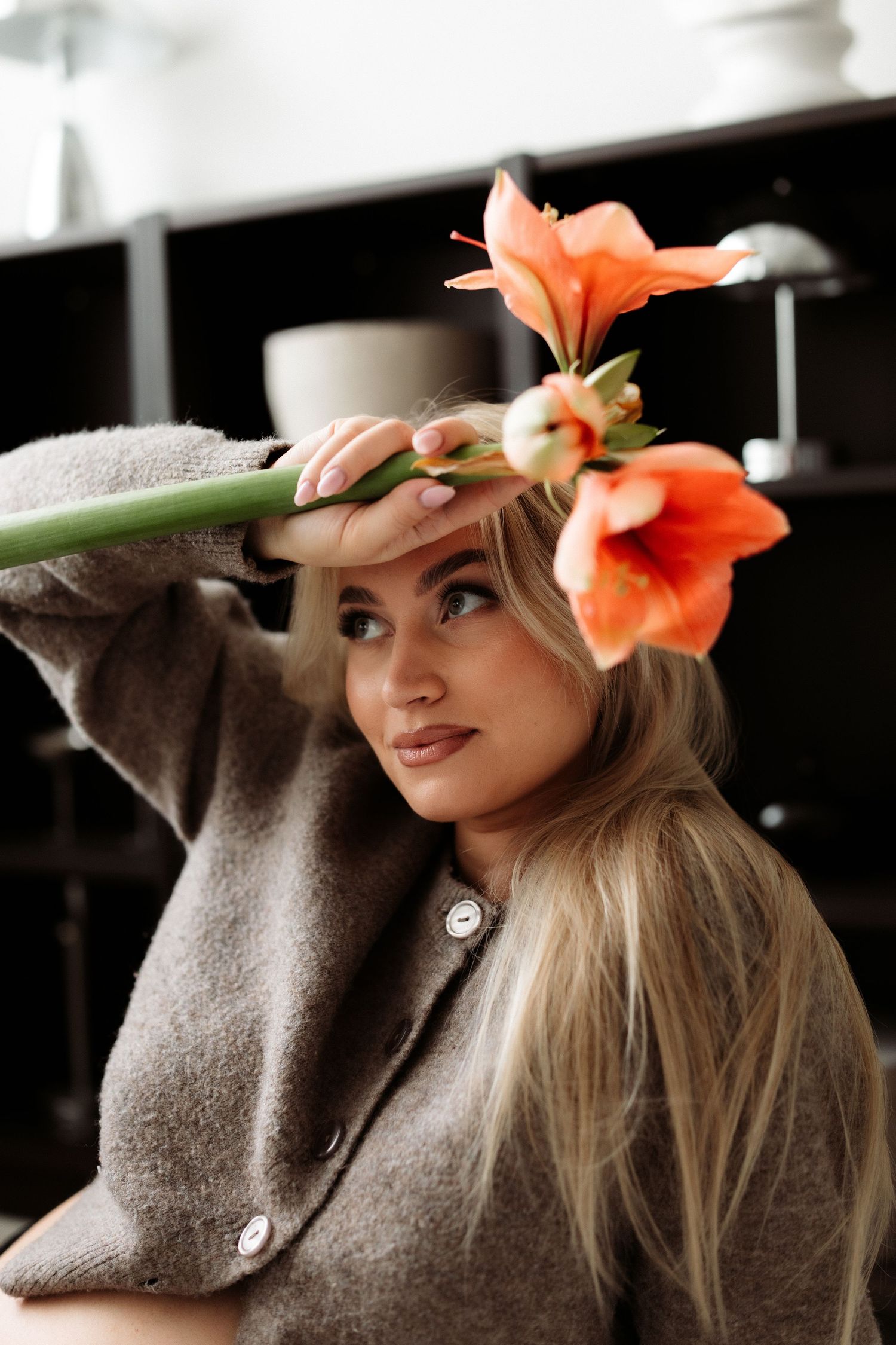 Blonde woman in brown cardigan holding orange amaryllis flowers near her face indoors.
