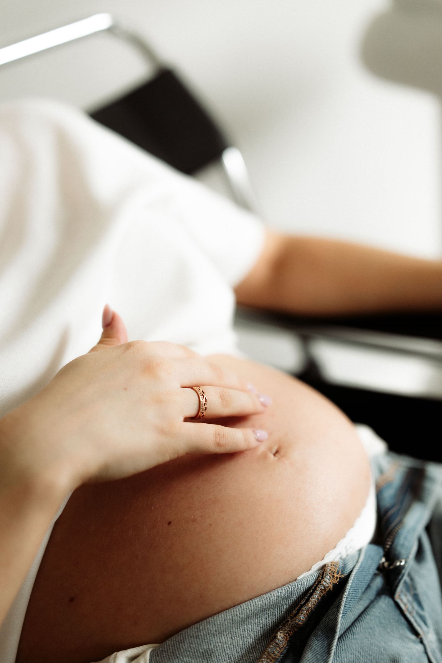 Pregnant woman in white shirt and jeans resting hand with gold ring on baby bump.