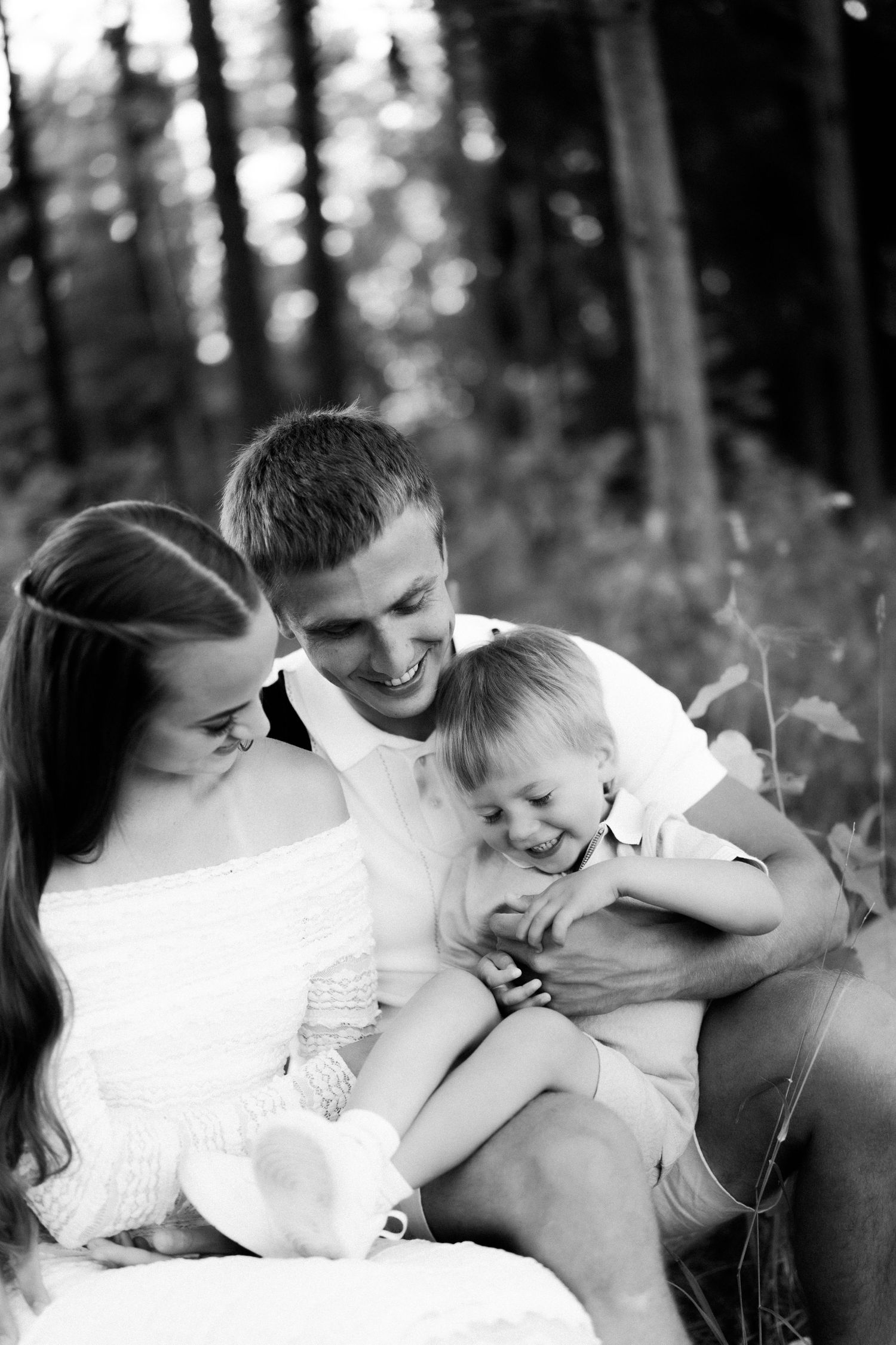 Black and white photo of happy young family with toddler laughing together outdoors in a forest setting.