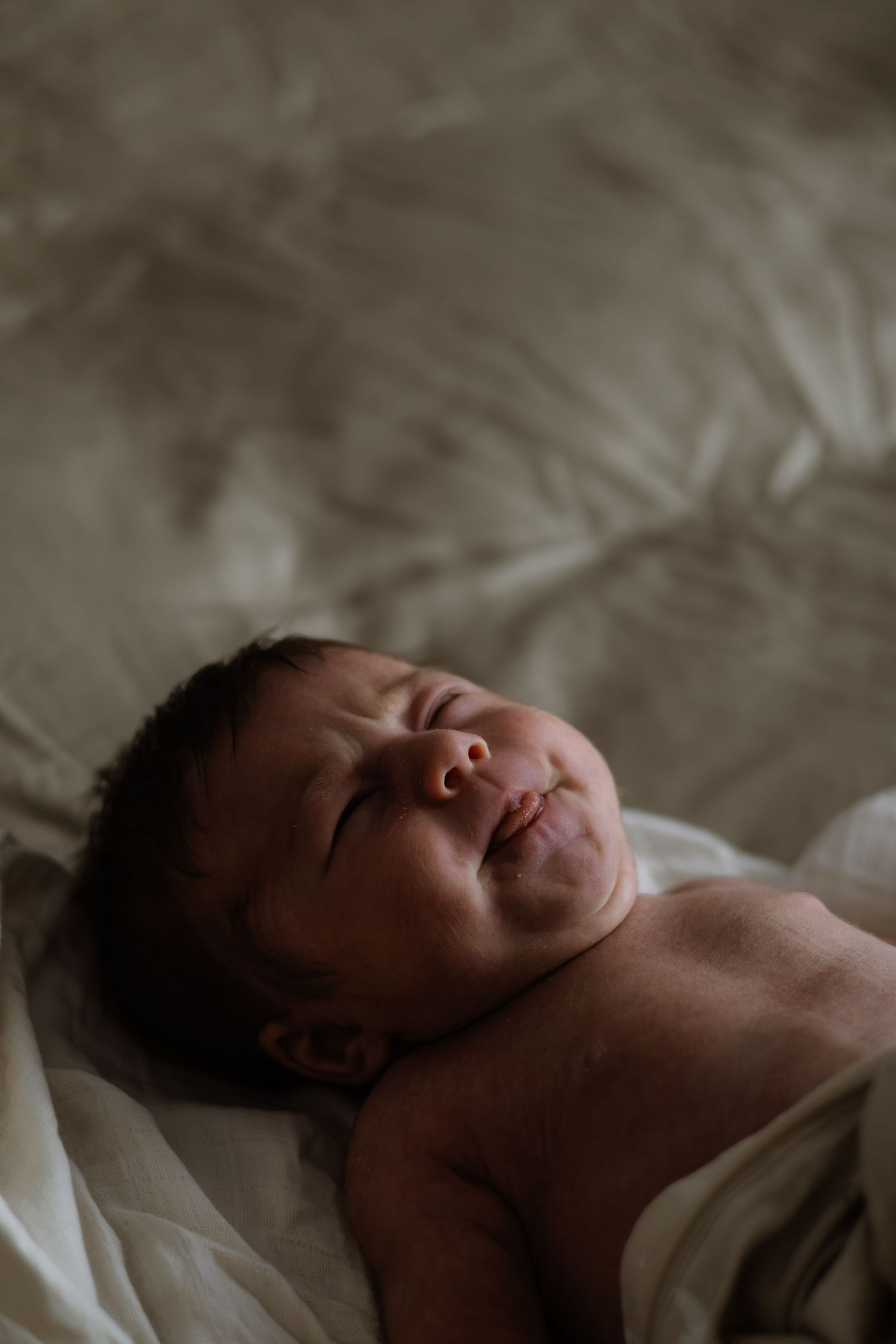 A peaceful newborn baby lying on white sheets, eyes closed, with dark hair and tiny features.