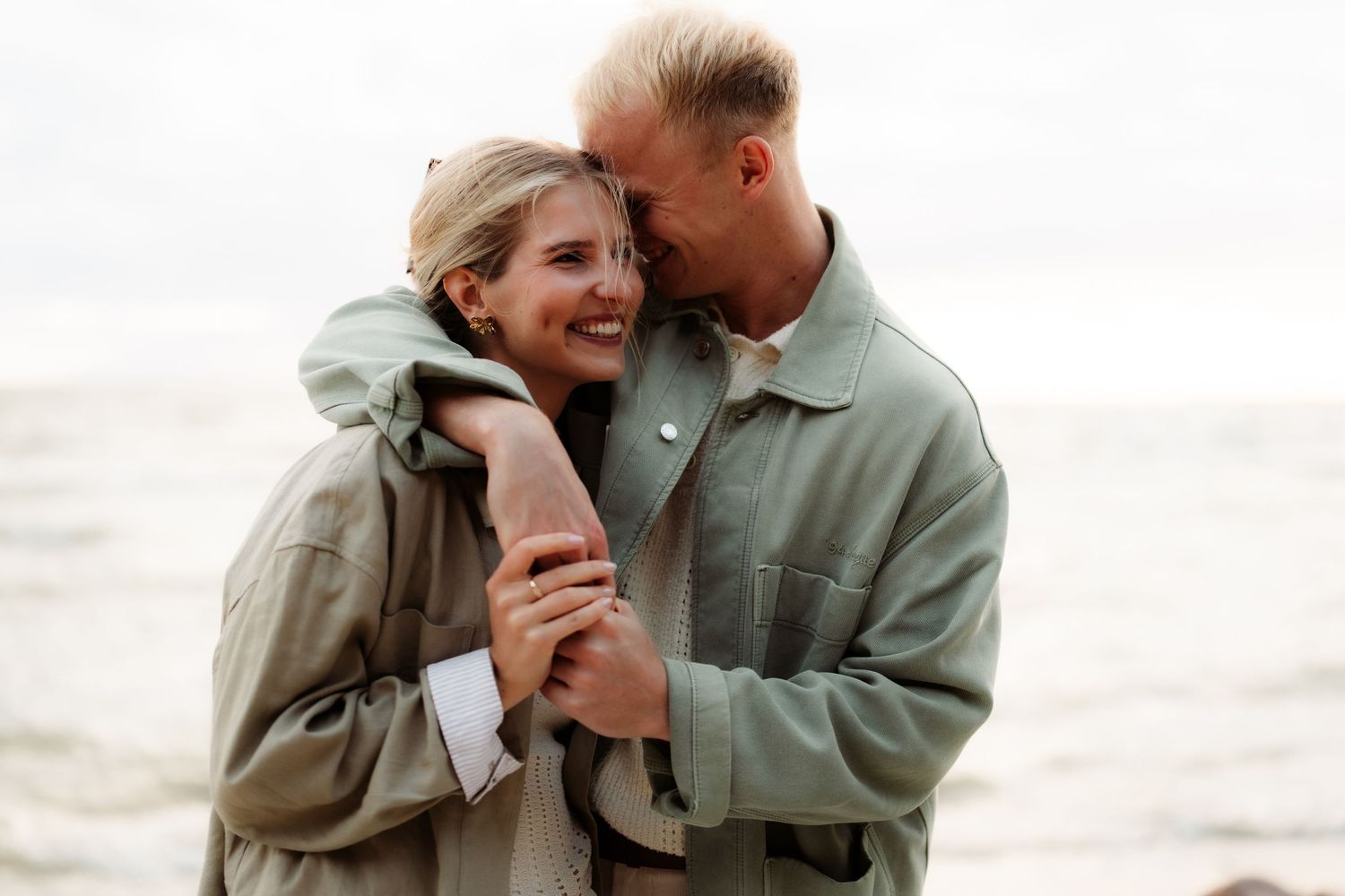 Happy couple embracing and smiling together on a cloudy beach, wearing matching sage green jackets.