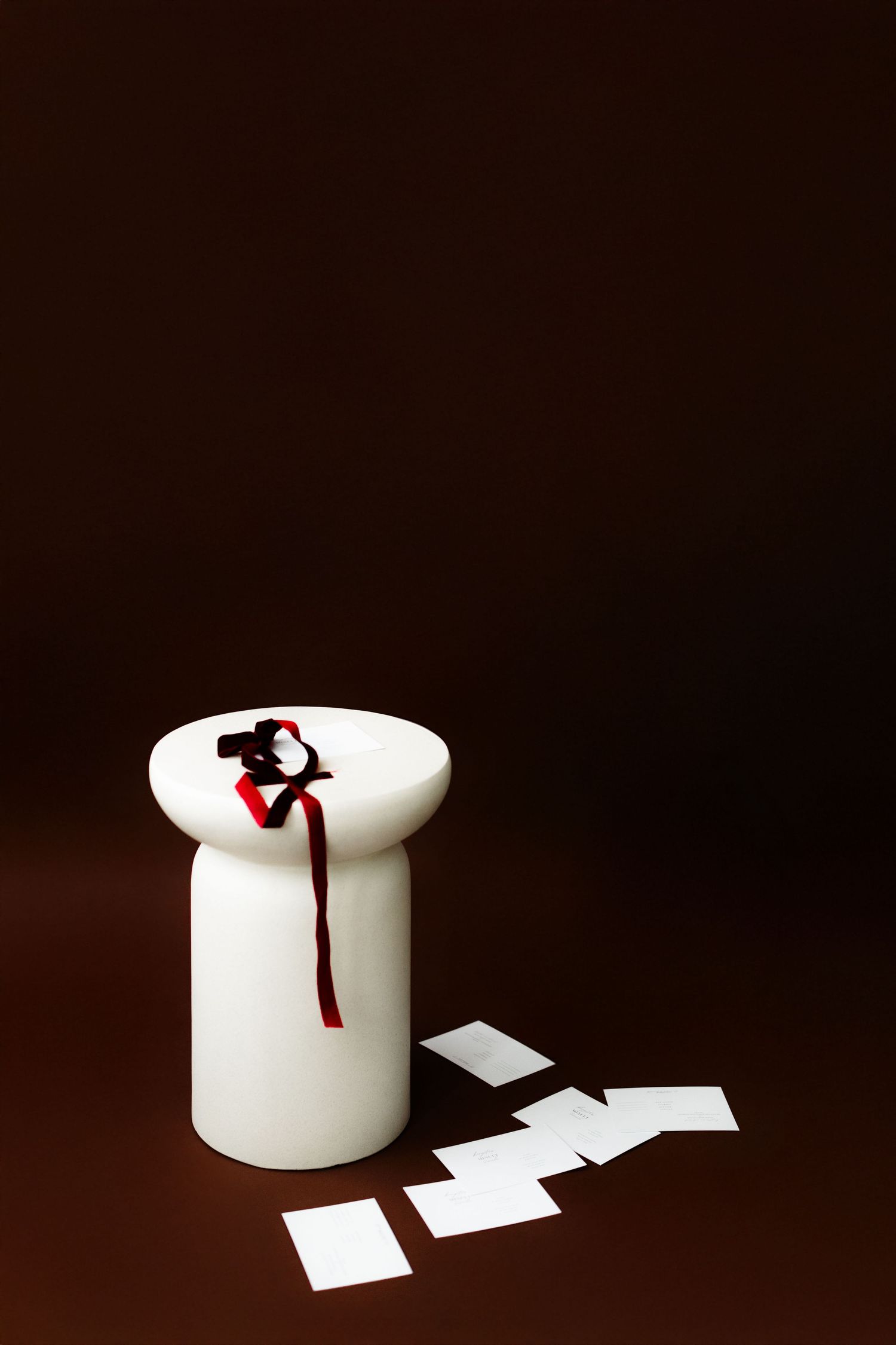 White ceramic jar with red ribbon and scattered paper pieces on dark brown background creates minimalist composition.