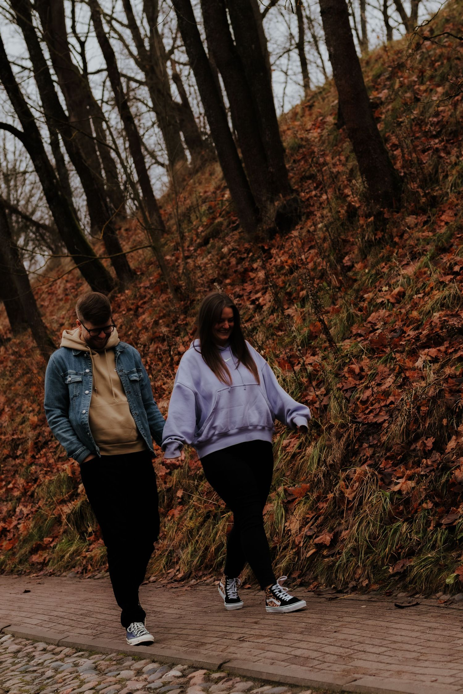 Two friends walking together on a brick path surrounded by autumn foliage and bare trees in a park setting.