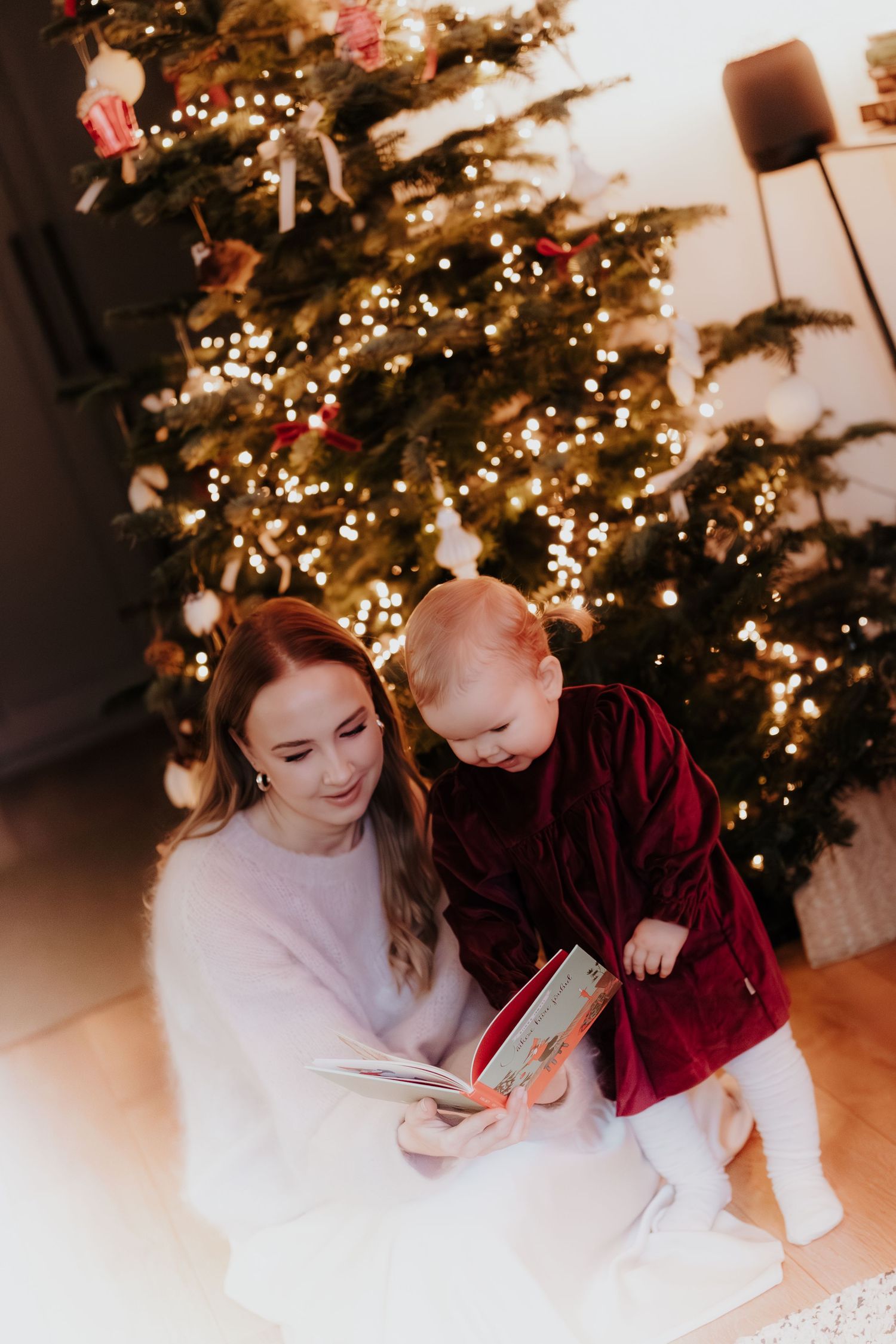Two people sit together on the floor reading by the warm glow of a decorated Christmas tree with twinkling lights.