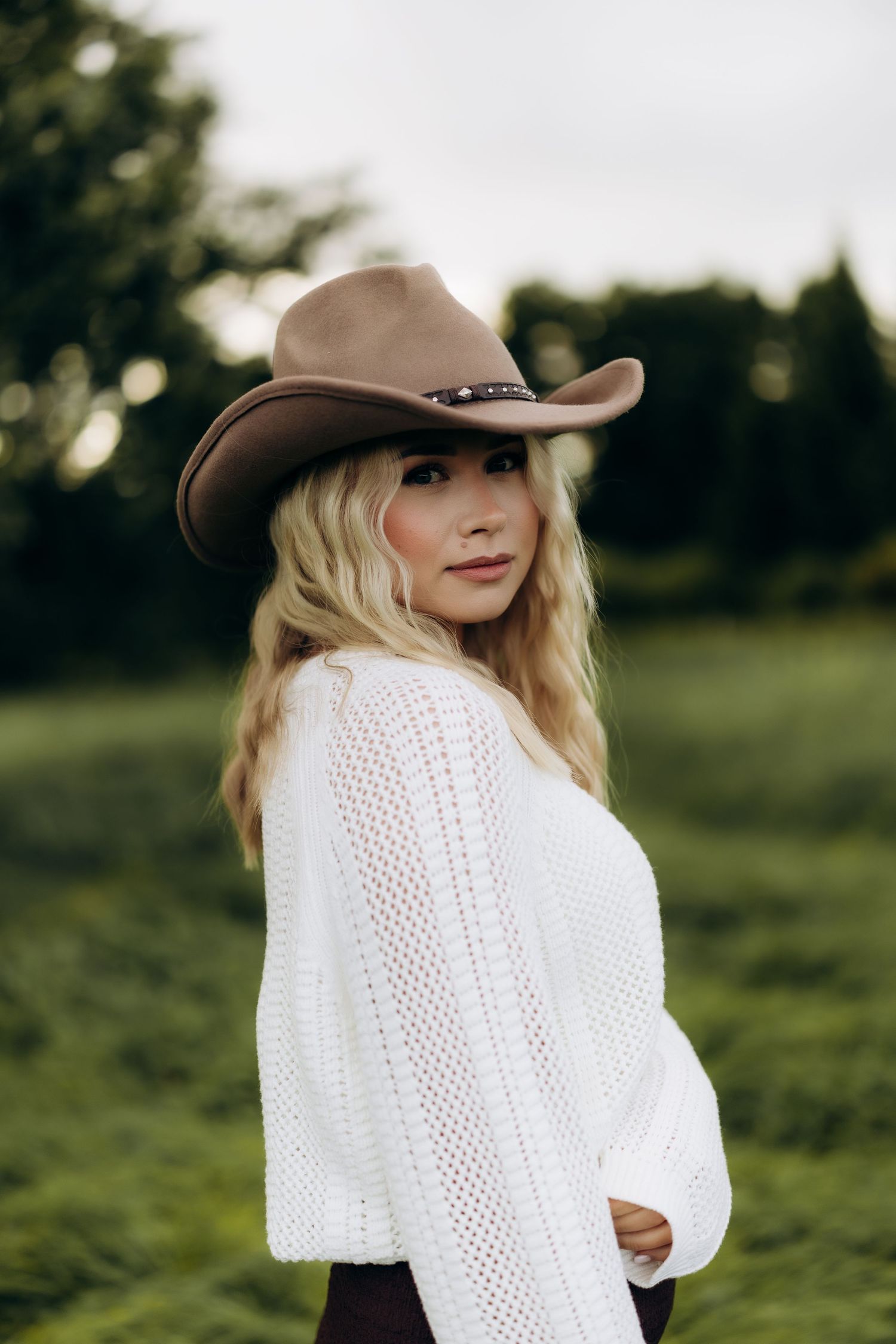 Blonde woman wearing a brown cowboy hat and white knit sweater posing outdoors in a green field.