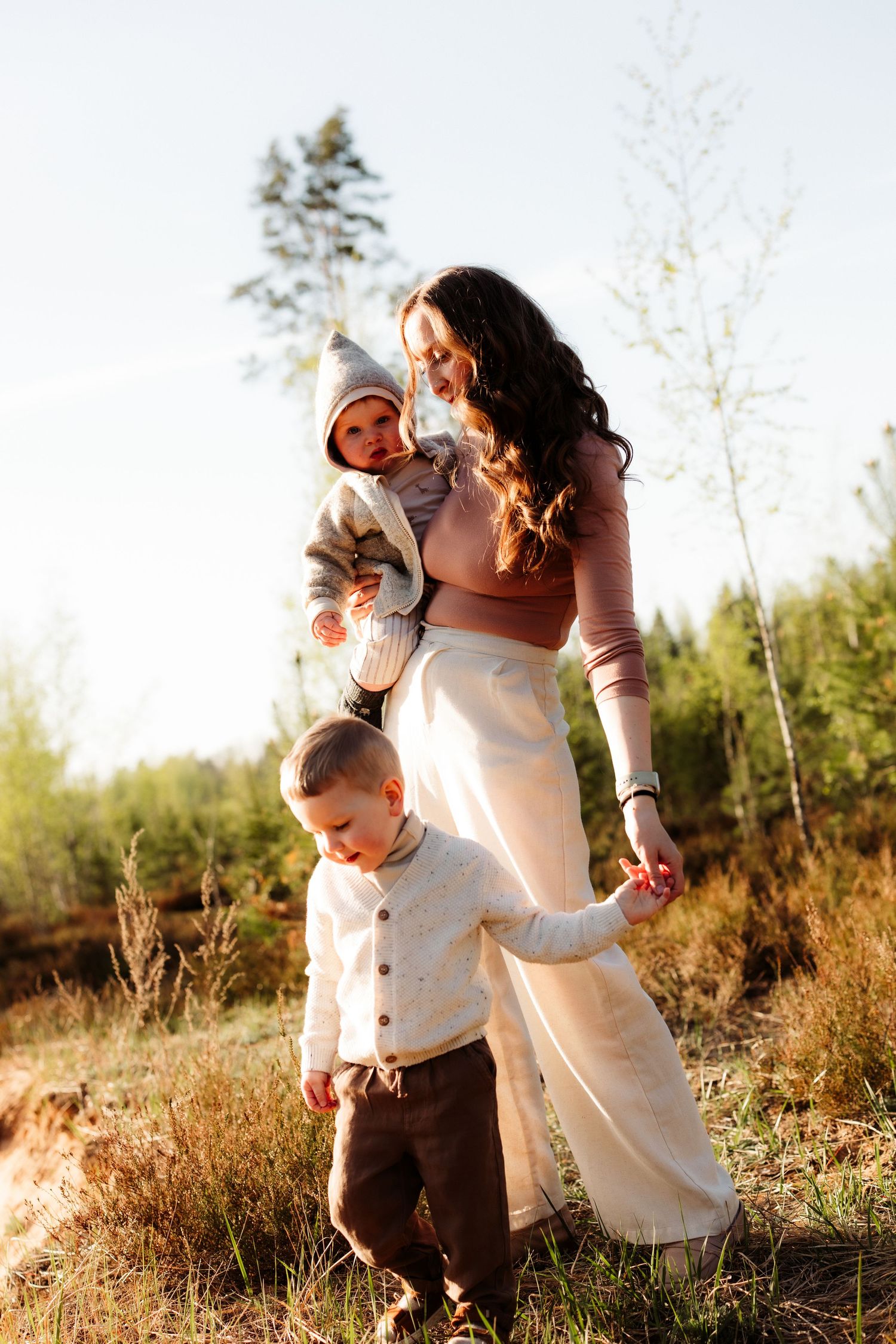 A young mother holds a baby while walking with a toddler outdoors in a sunlit forest clearing.