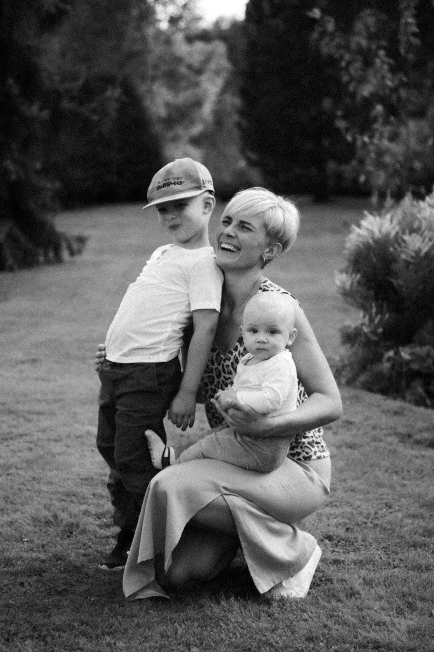 A black and white portrait of a parent and two children sharing a joyful moment together on a grassy lawn.