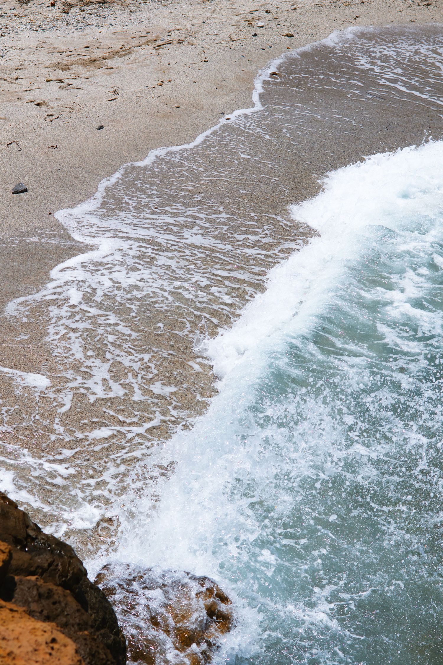 Aerial view of turquoise ocean waves crashing against a sandy beach with rocky cliffs visible in the foreground.