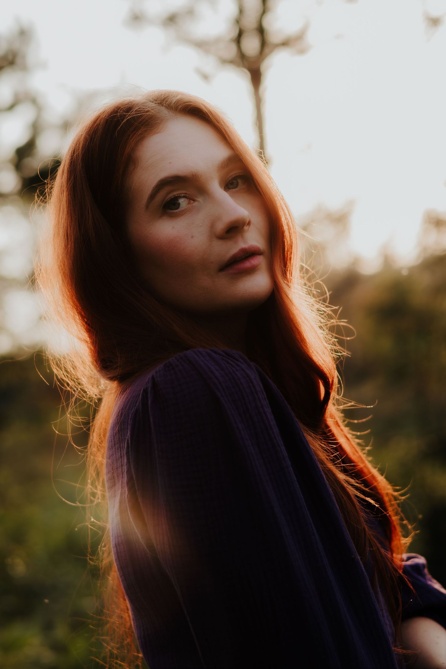 A woman with flowing red hair gazes upward in golden sunset light while wearing a navy blue sweater.