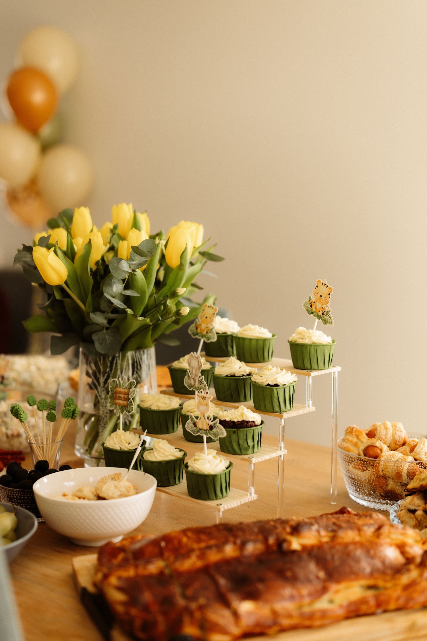 A party buffet table with yellow tulips, tiered cupcakes, and savory dishes including a golden brown pastry.