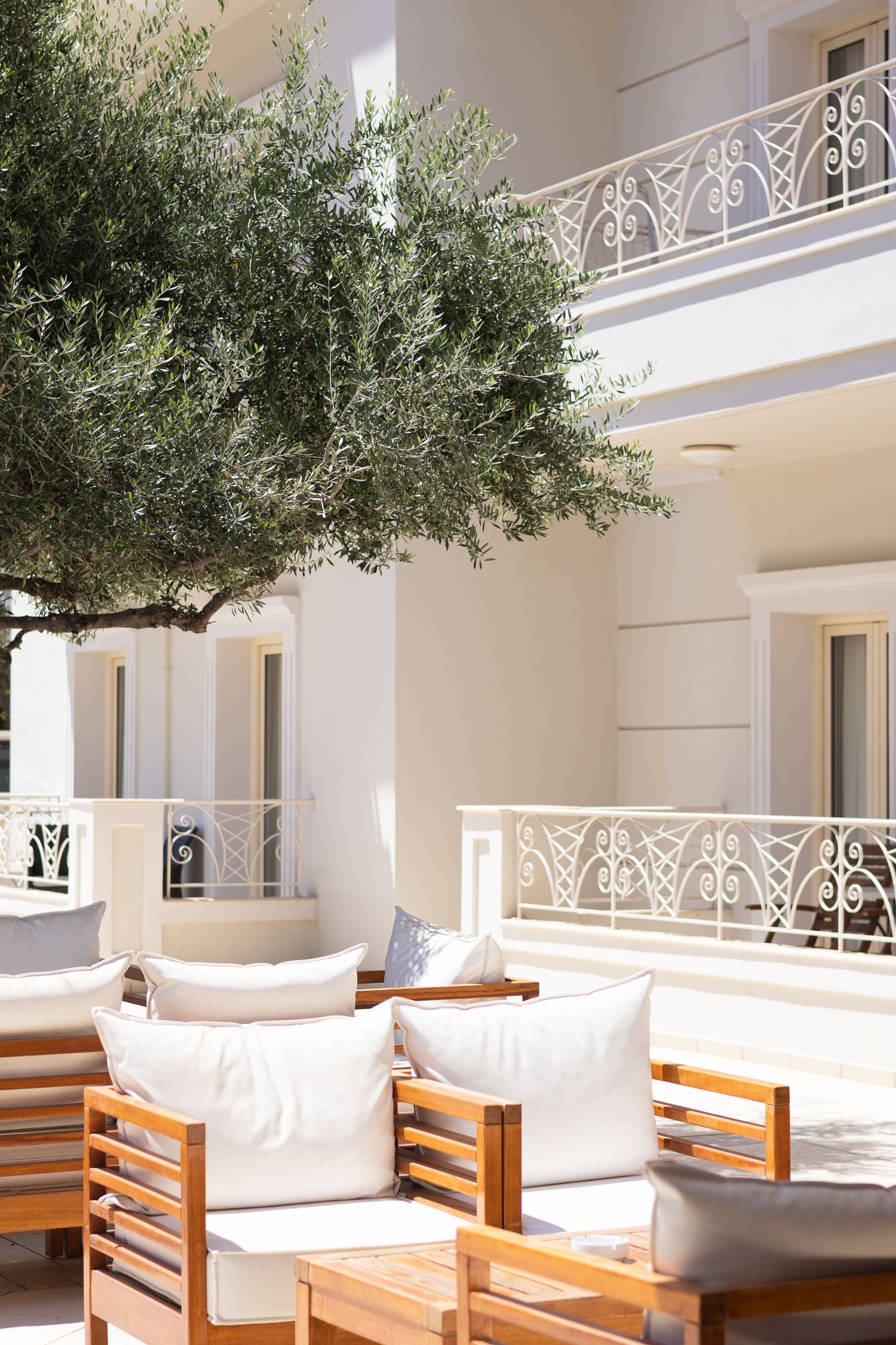 Elegant Mediterranean patio with olive trees, white railings, and teak outdoor furniture with plush cushions.