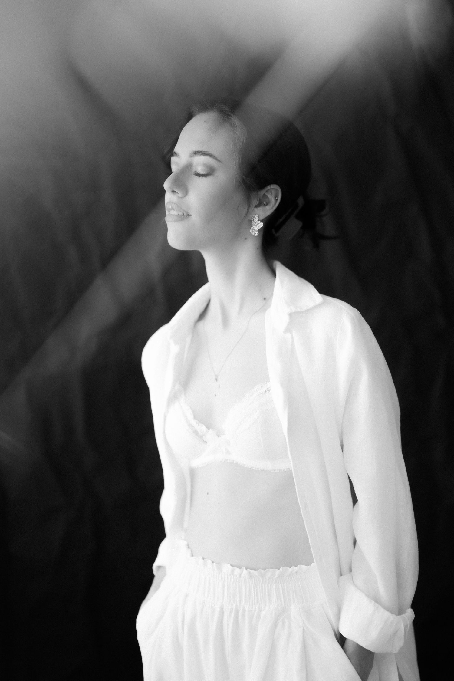 Black and white portrait of a woman in white blazer and bra top, eyes closed, bathed in dramatic light rays.
