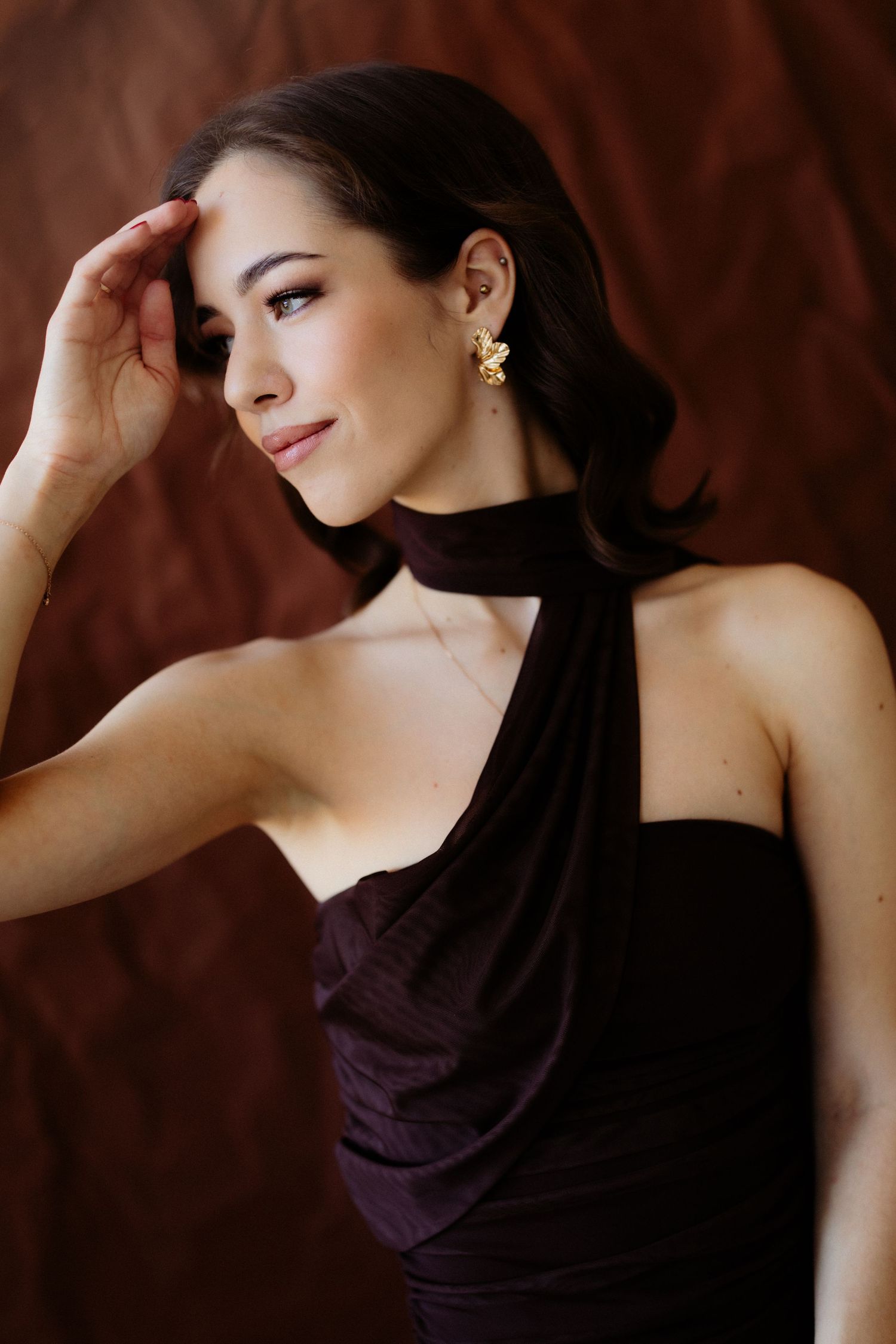 Elegant woman in dark purple halter dress with gold earrings posing against a rich brown backdrop.