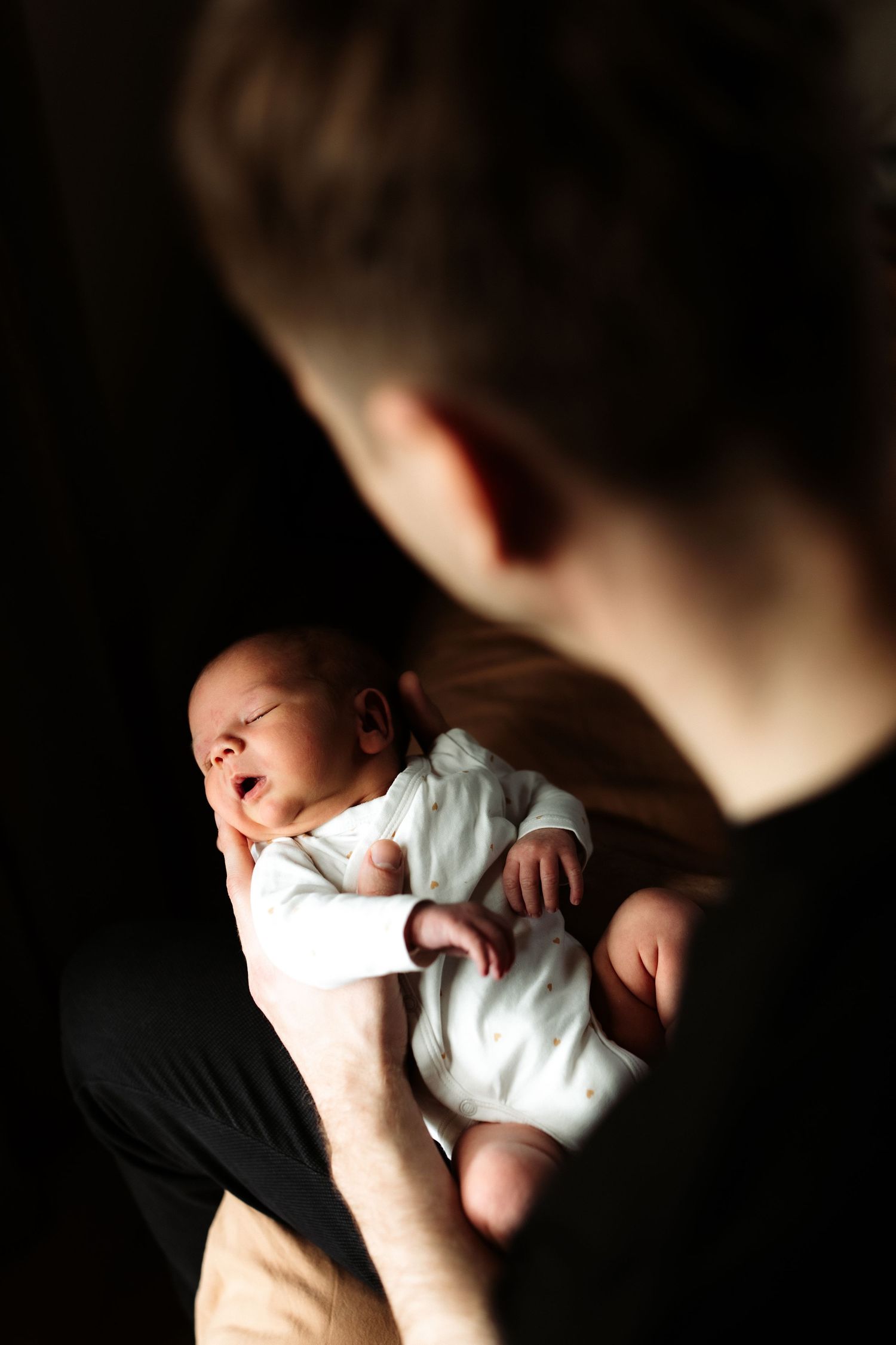 A father tenderly holds his newborn baby in both hands against a dark background.