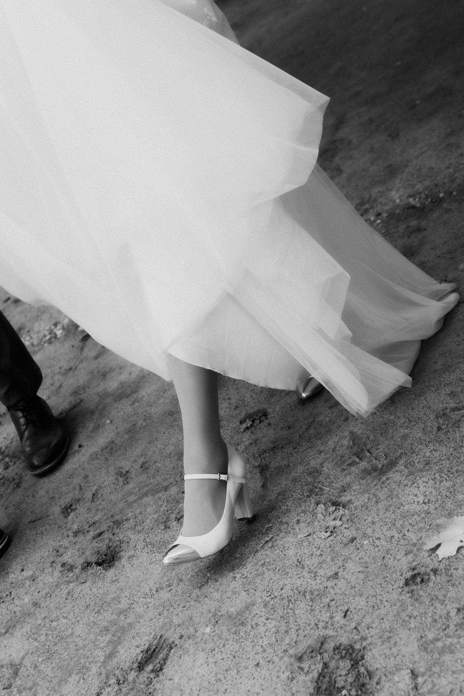 A close-up black and white photo of white heels and flowing tulle dress fabric against a textured ground.