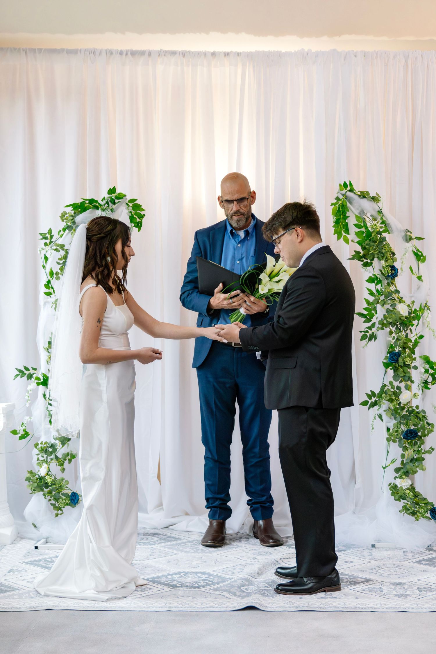 Bride places ring on groom's finger during indoor wedding ceremony before white drape backdrop with green garlands.