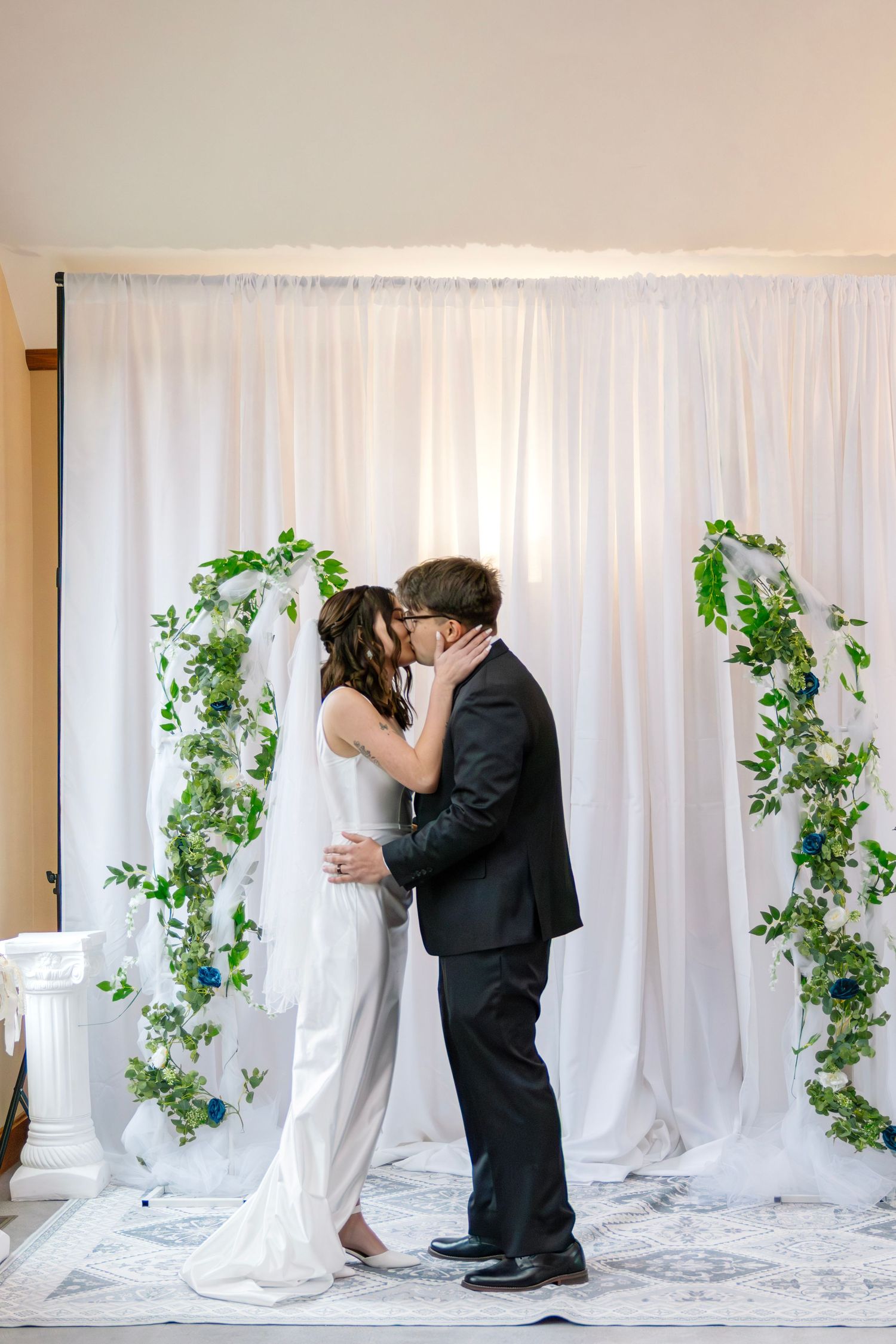 Newlyweds share their first kiss at the altar before a white draped backdrop with lush green floral garlands.