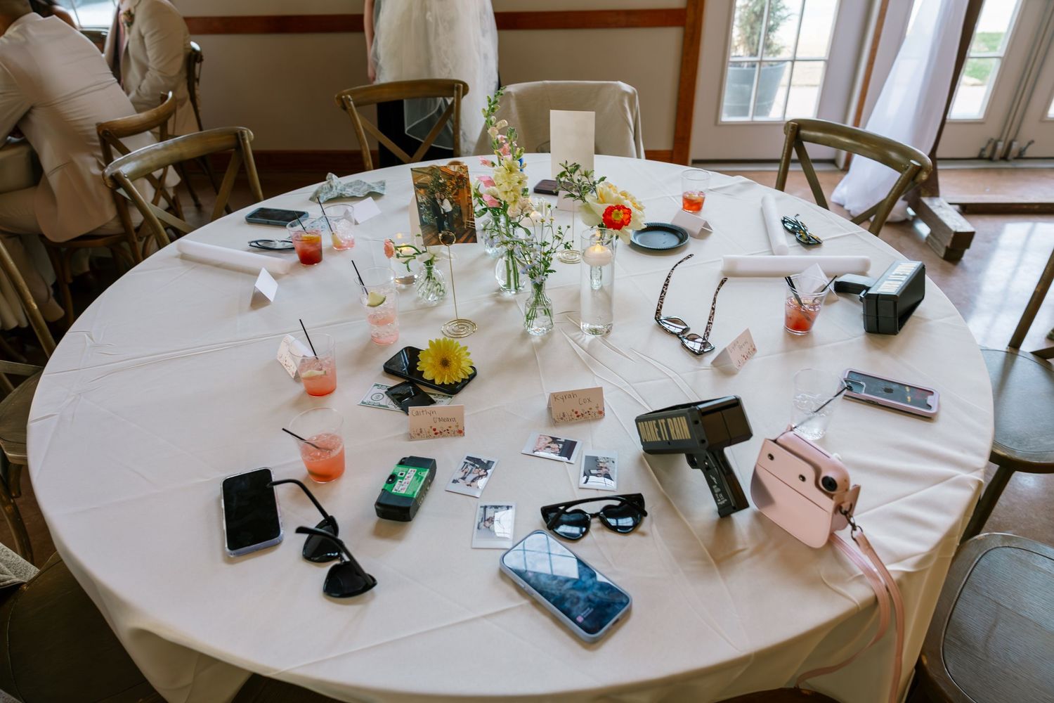 Round wedding reception table with white linen covered in phones, sunglasses, cameras, and floral centerpieces.