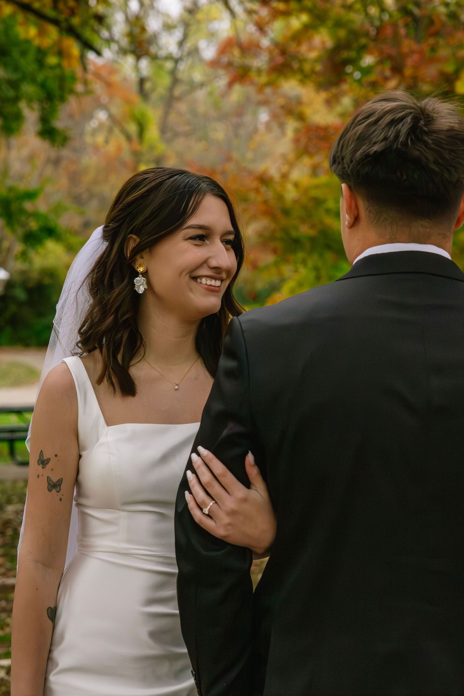 Smiling bride in white dress and veil with butterfly tattoo holds groom's arm outdoors in autumn park.