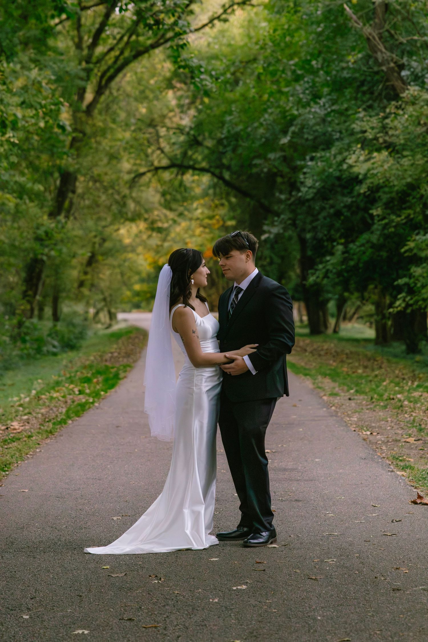 Bride and groom face each other holding hands on tree-lined park path in elegant wedding attire.