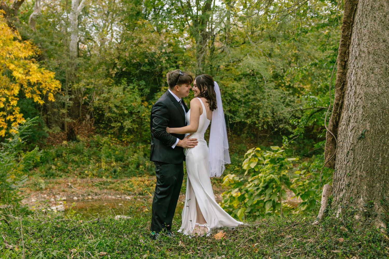 Bride and groom share romantic moment kissing in lush green forest setting during fall wedding.